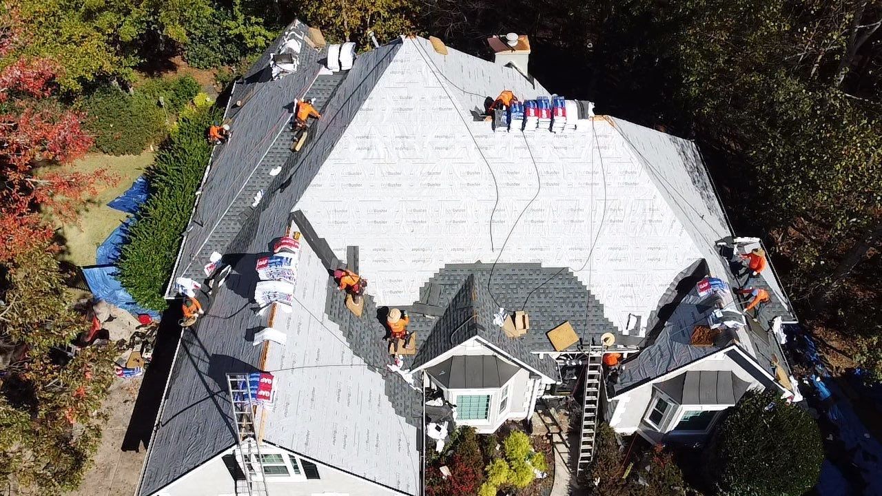 Workers installing shingles on a multi-sectioned roof; trees surround the house.