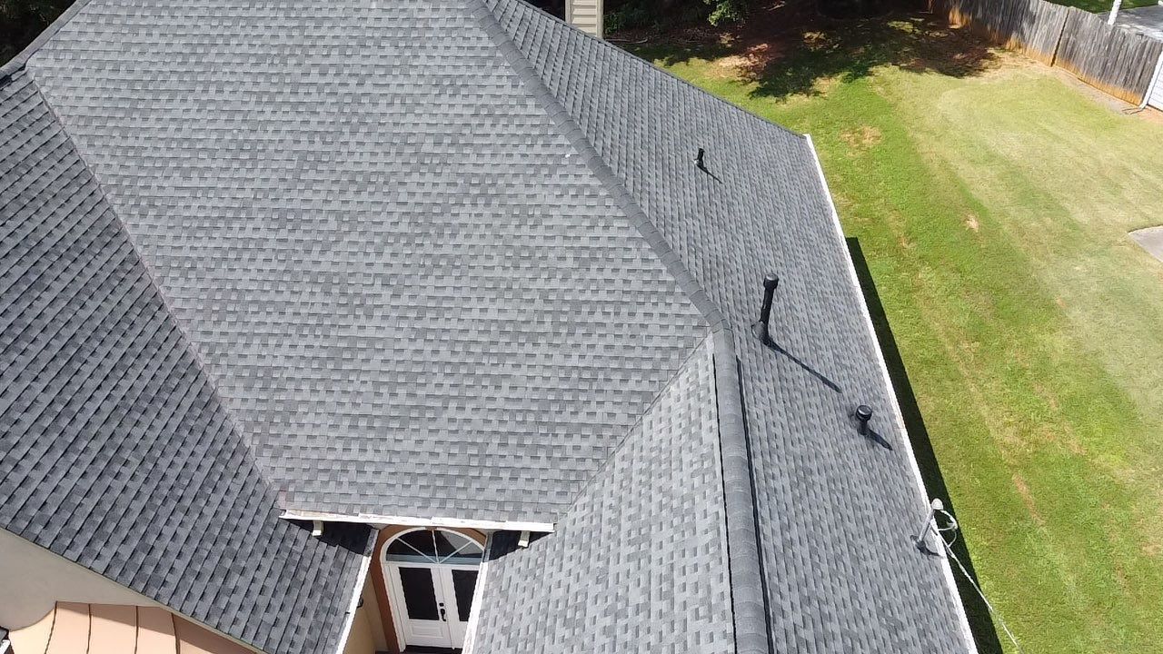 Gray shingled roof of a house, with a chimney and part of a dormer. Green lawn visible.