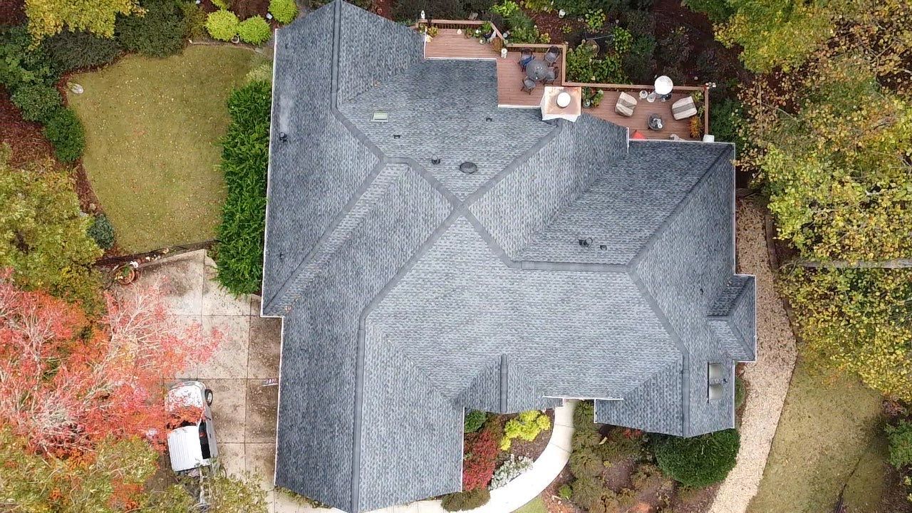 Overhead view of a gray shingled roof with a deck, surrounded by a yard with trees.