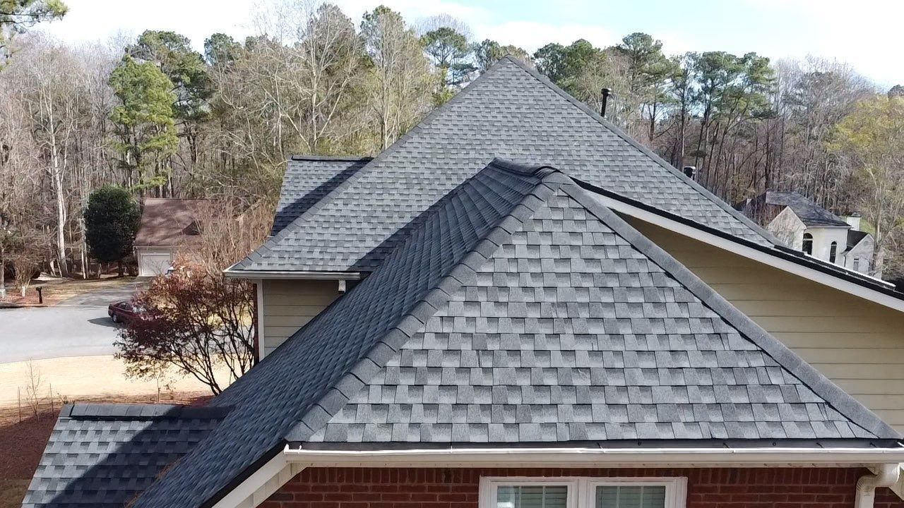 Gray asphalt shingle roof on a beige house with brick trim, set against a background of trees.