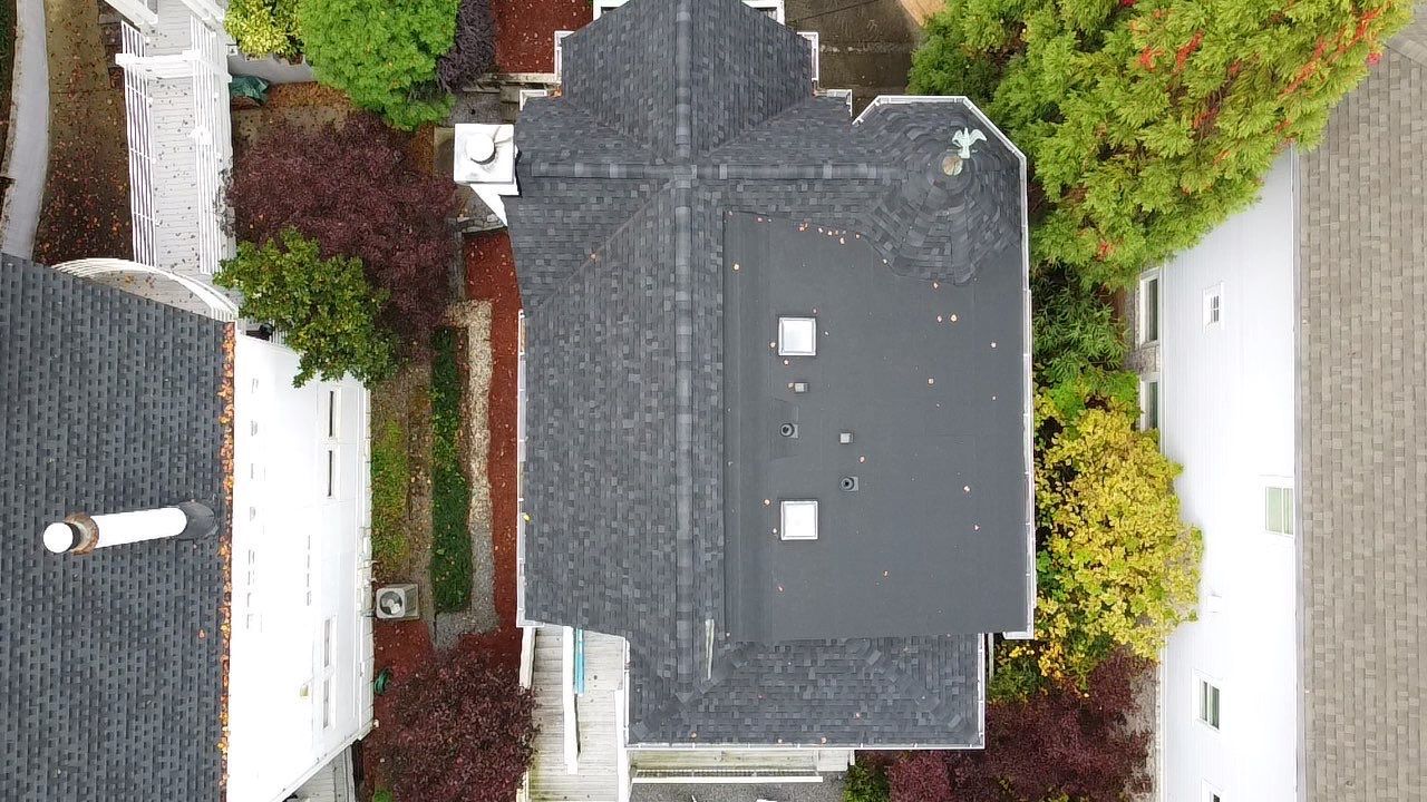 Overhead view of a house with a dark gray roof surrounded by trees and neighboring houses.