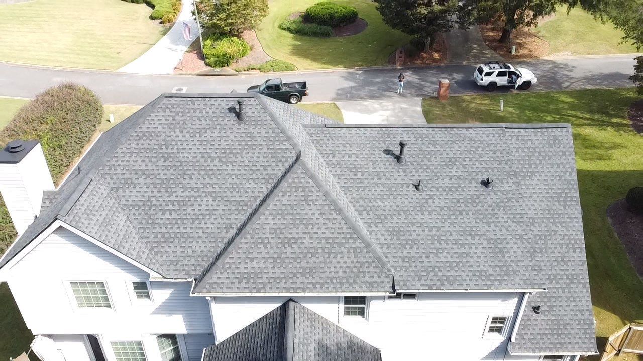 Overhead view of a house with a gray shingled roof, trees, and a road.