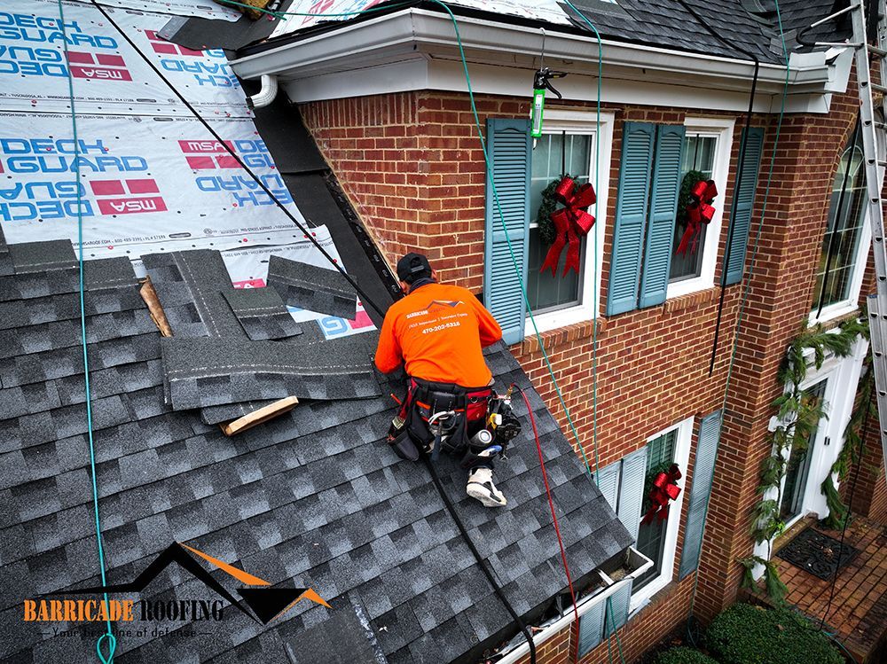 Roofer in orange shirt on a roof, installing shingles, with safety harness, near a brick building.