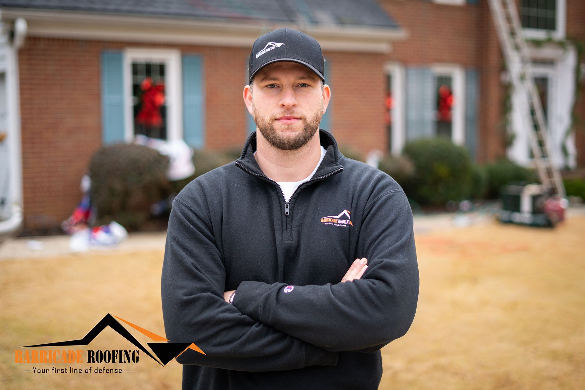 Man in black sweatshirt and cap with arms crossed, in front of a brick house, logo in lower left corner.