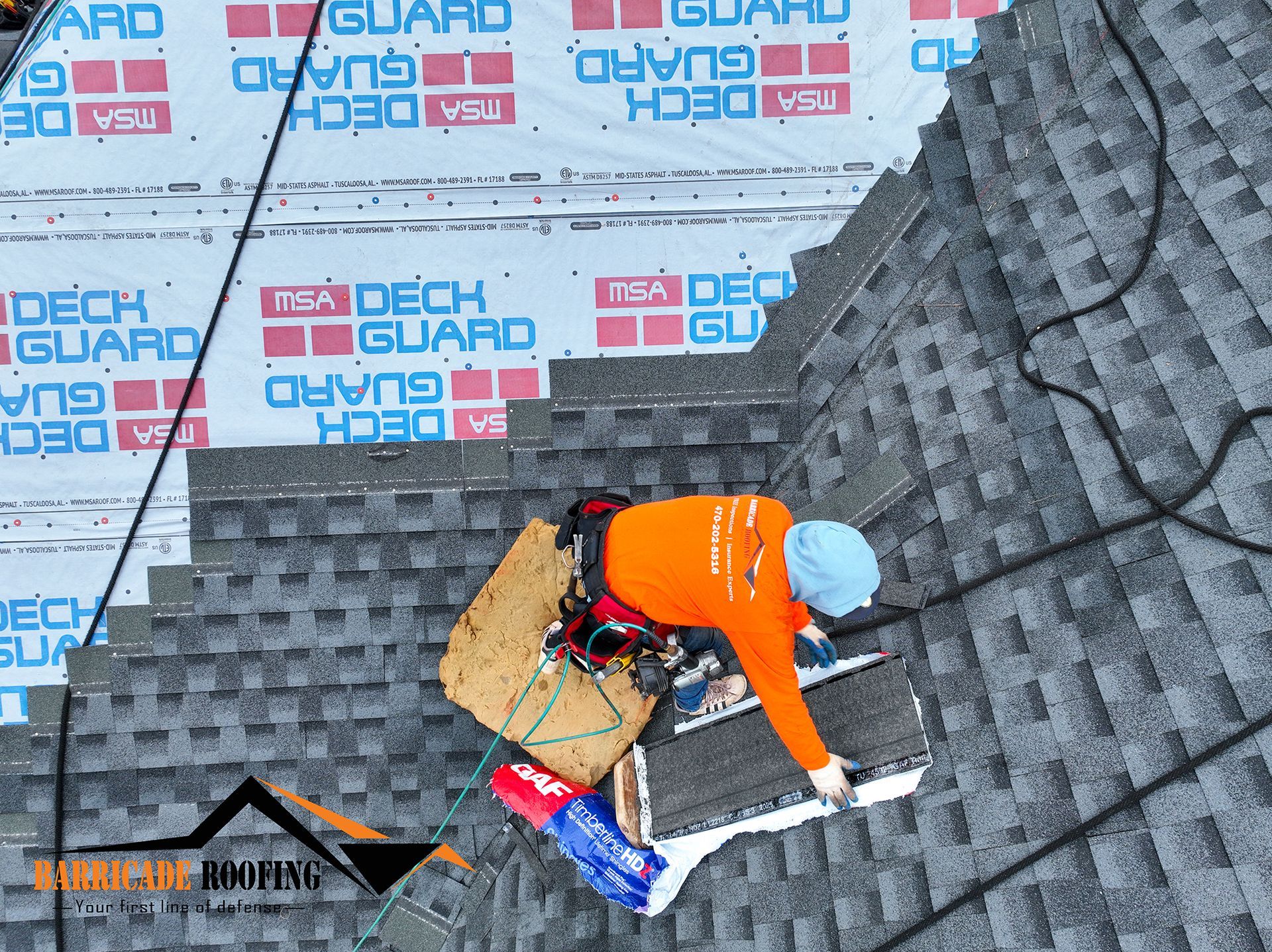 Roofer in orange shirt, installing shingles. Safety harness in use. Deck Guard underlayment visible.