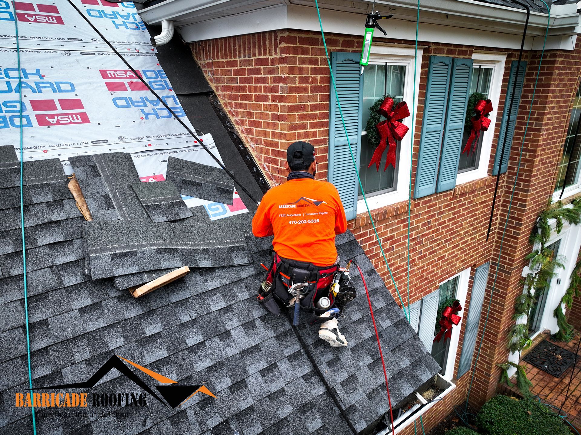 Roofer in orange shirt working on a shingled roof, near a brick house with Christmas decorations.