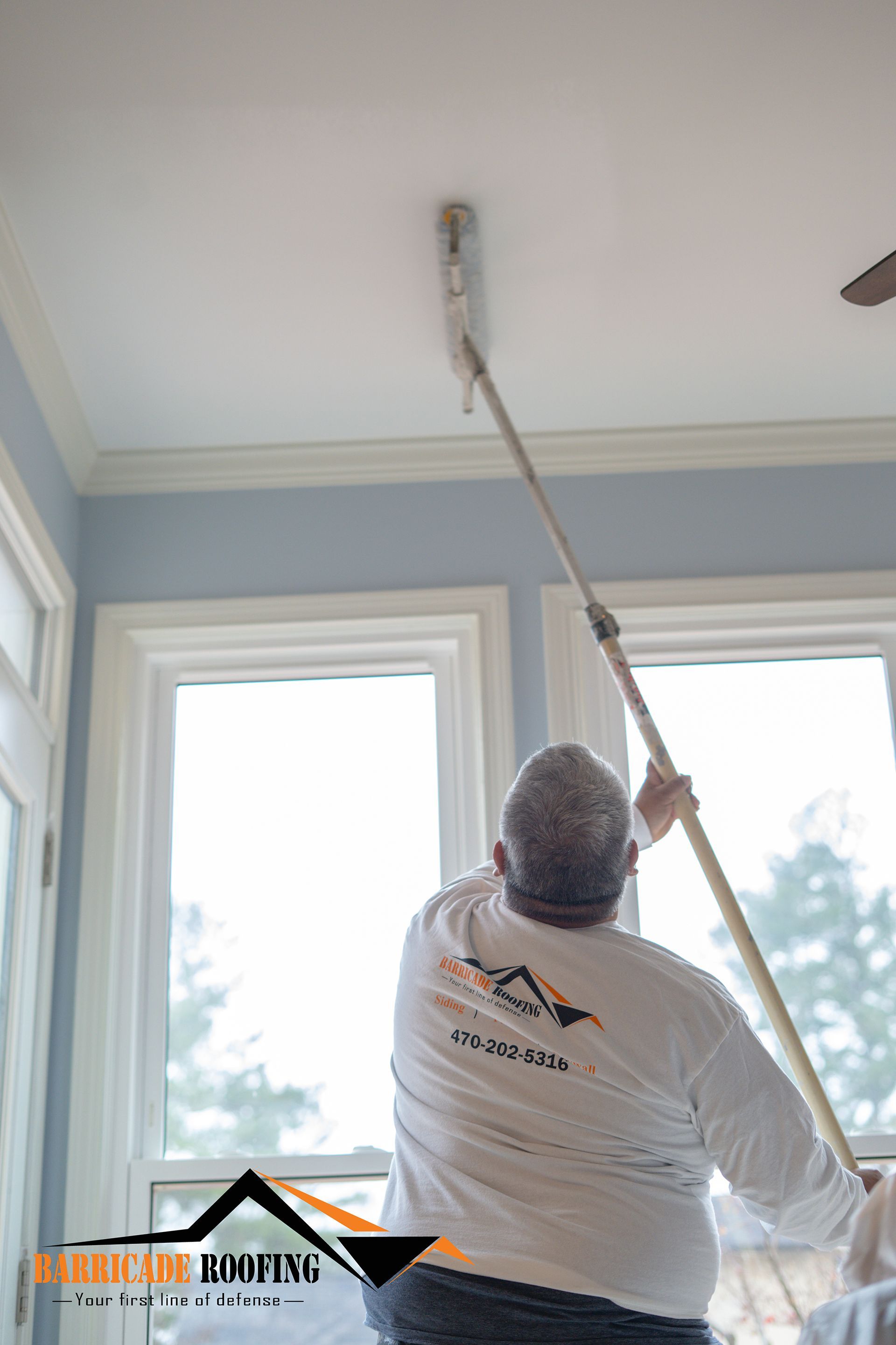Person painting a ceiling with a roller, in a room with white trim and large windows.