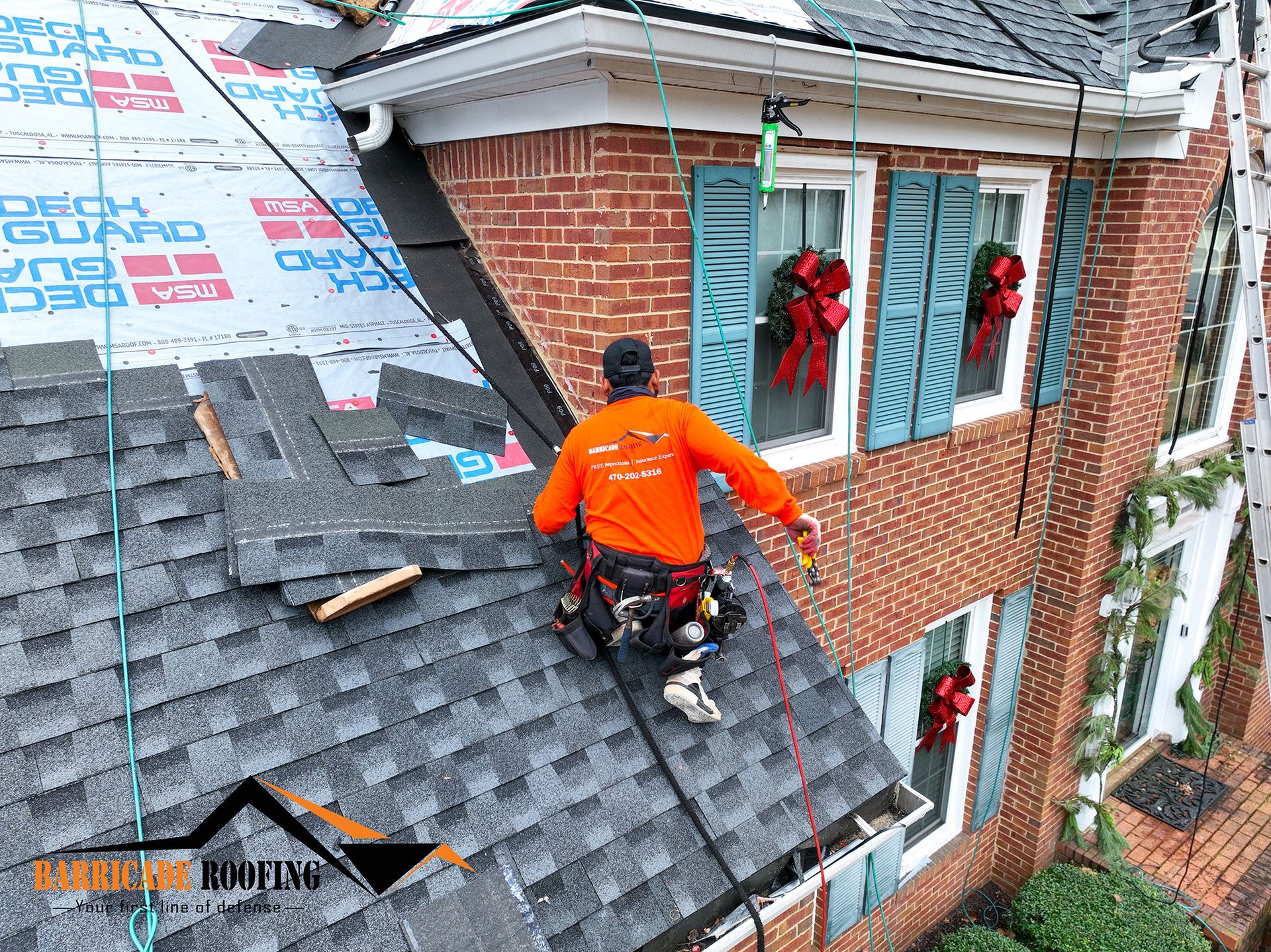 Roofer in an orange shirt working on a roof, attached to safety lines. Brick house with green shutters.