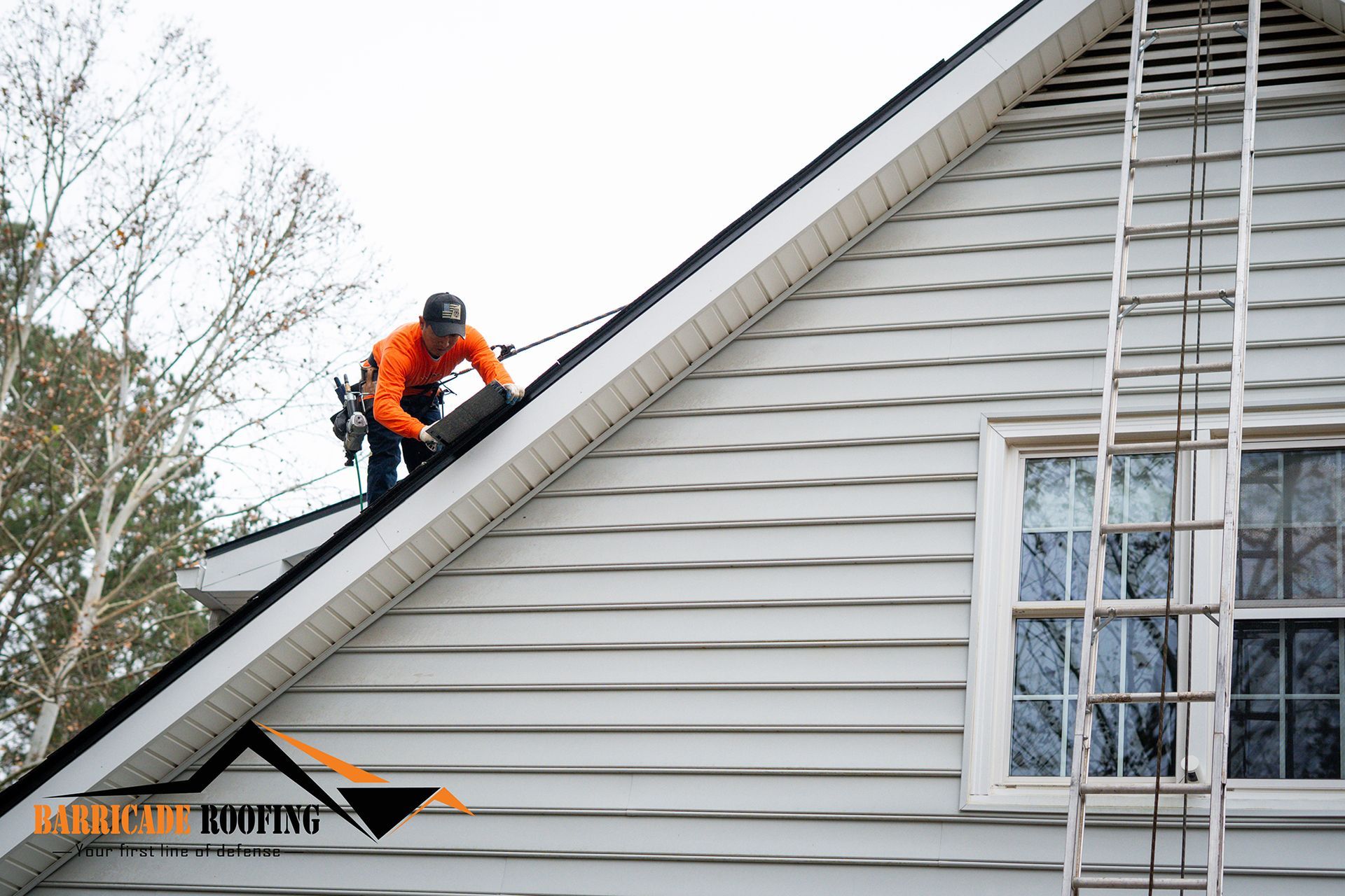 Roofer repairing a roof, secured by a safety harness. A-frame house with white siding and a ladder.