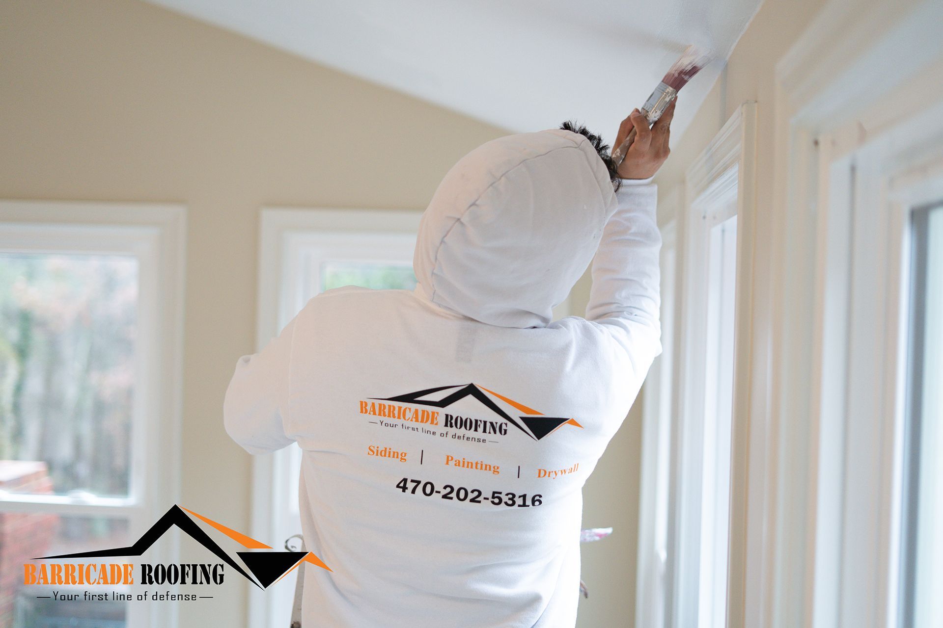 Person painting trim indoors. Wearing a hooded white sweatshirt with a company logo. Windows in the background.