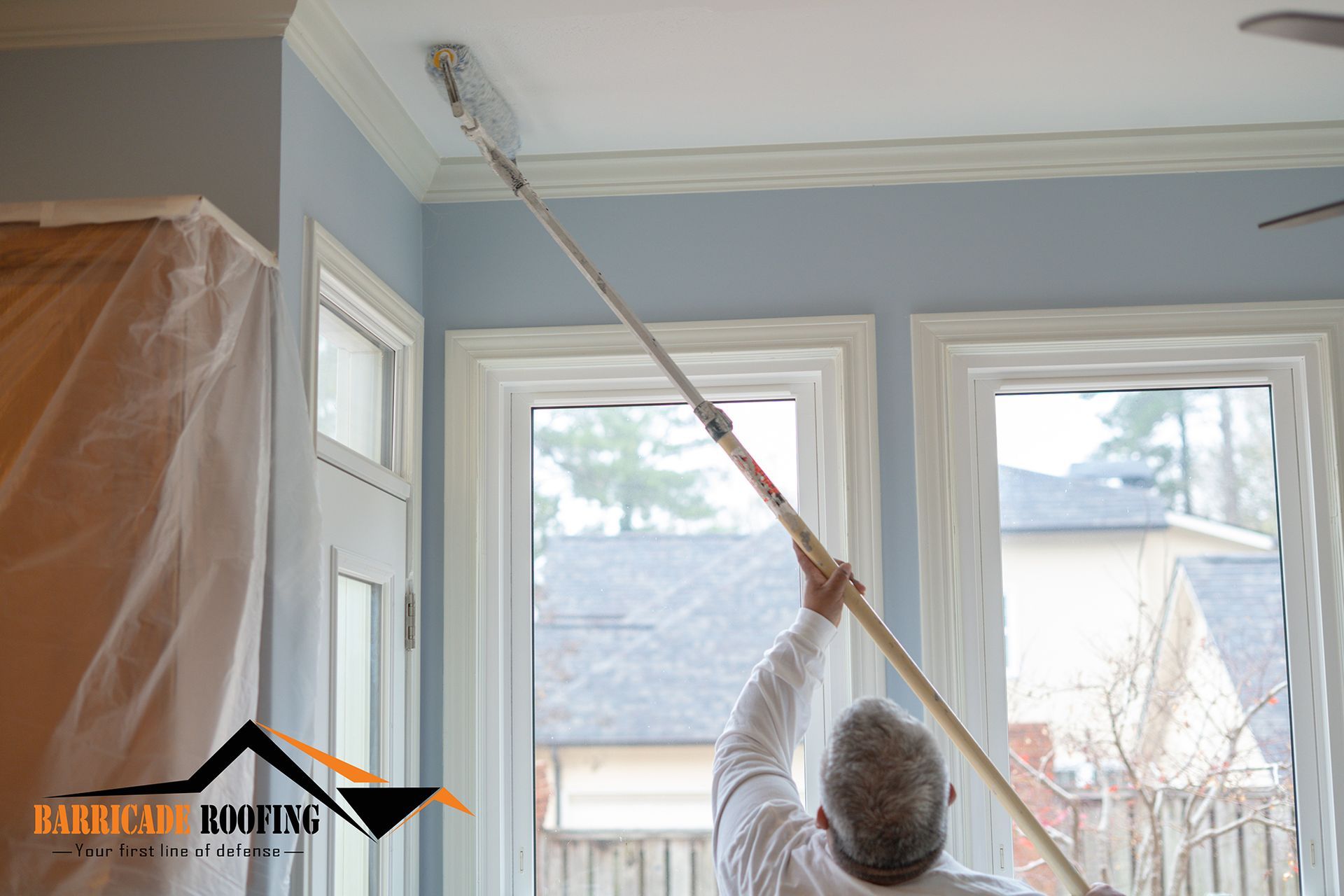 Person paints ceiling trim with a roller on an extension pole in a room with blue walls, windows, and white trim.