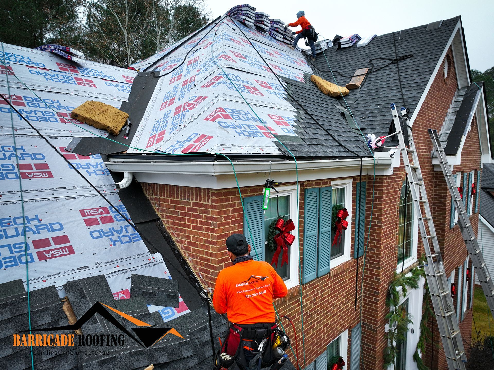 Roofers replacing shingles on a brick house; workers wearing safety gear; blue shutters.