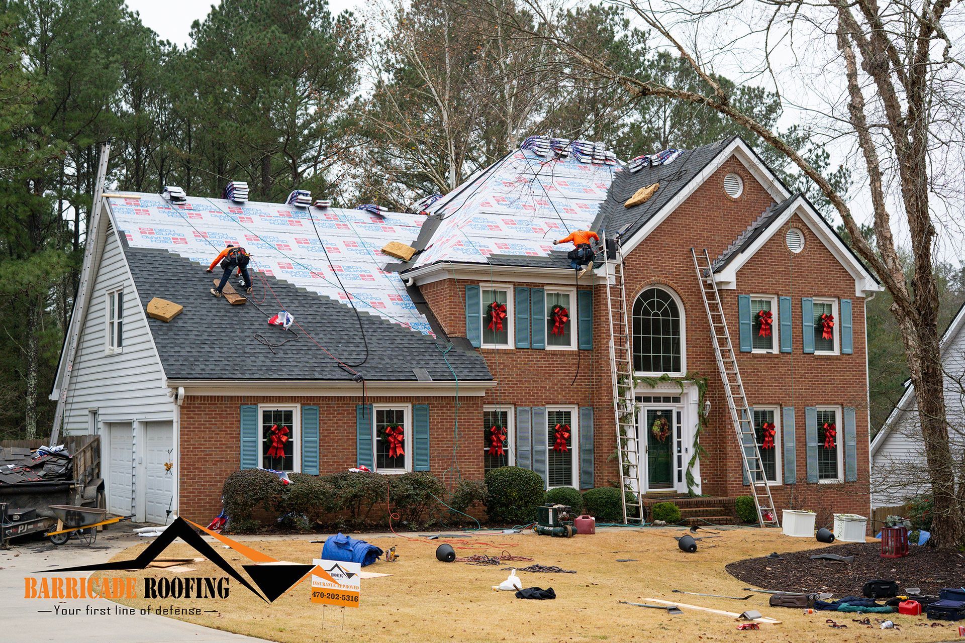 Roofers working on a two-story brick house with a new roof partially installed. Tools and materials on the ground.
