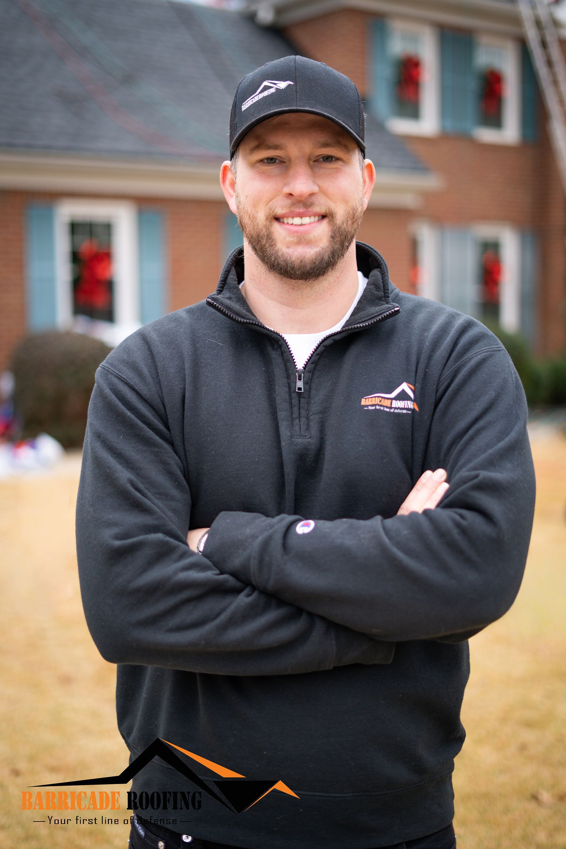 Man in black baseball cap and sweater, arms crossed, smiling in front of a house.