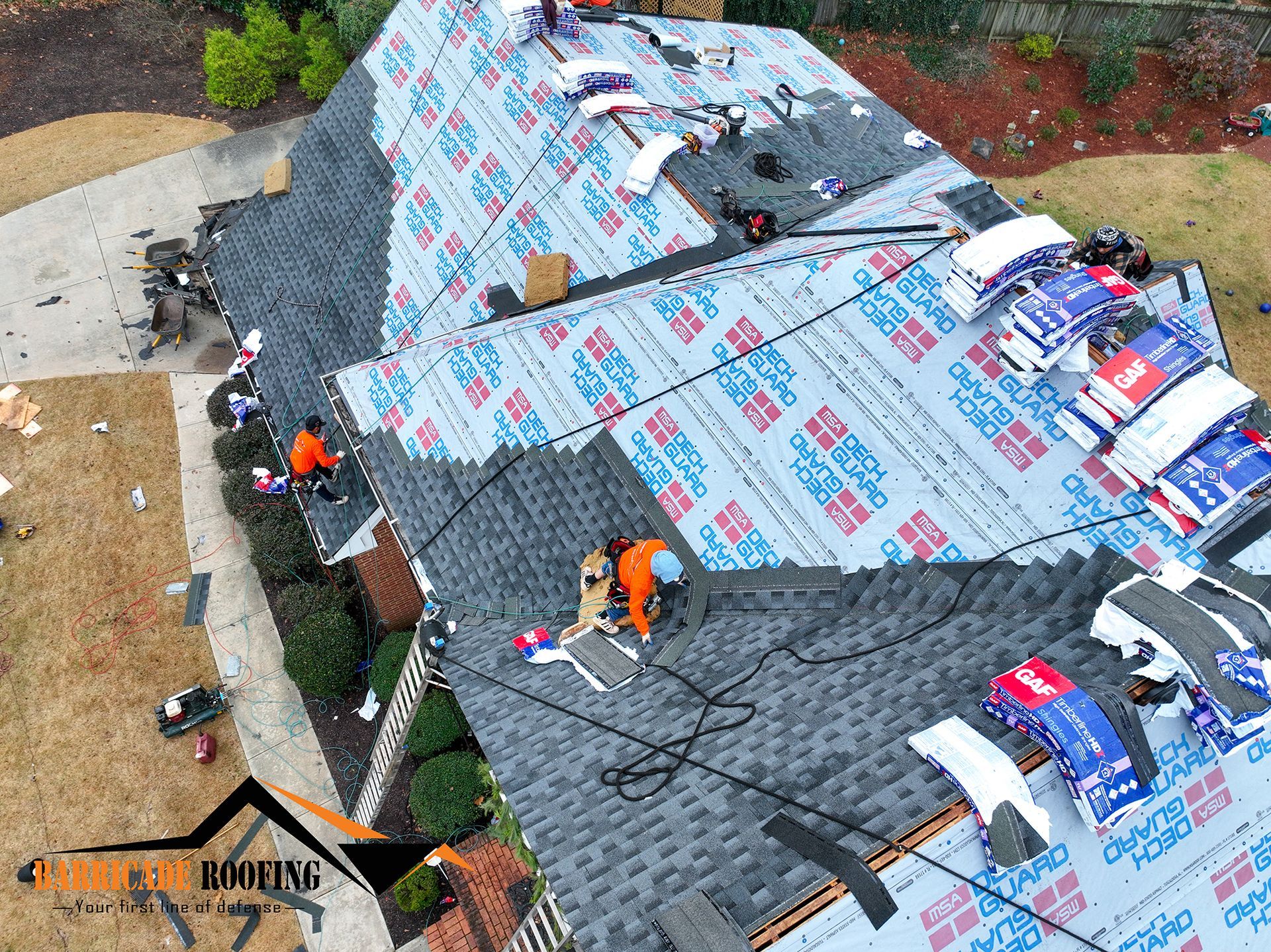 Roofers working on a house, partially covered in blue underlayment and shingles; supplies are present.