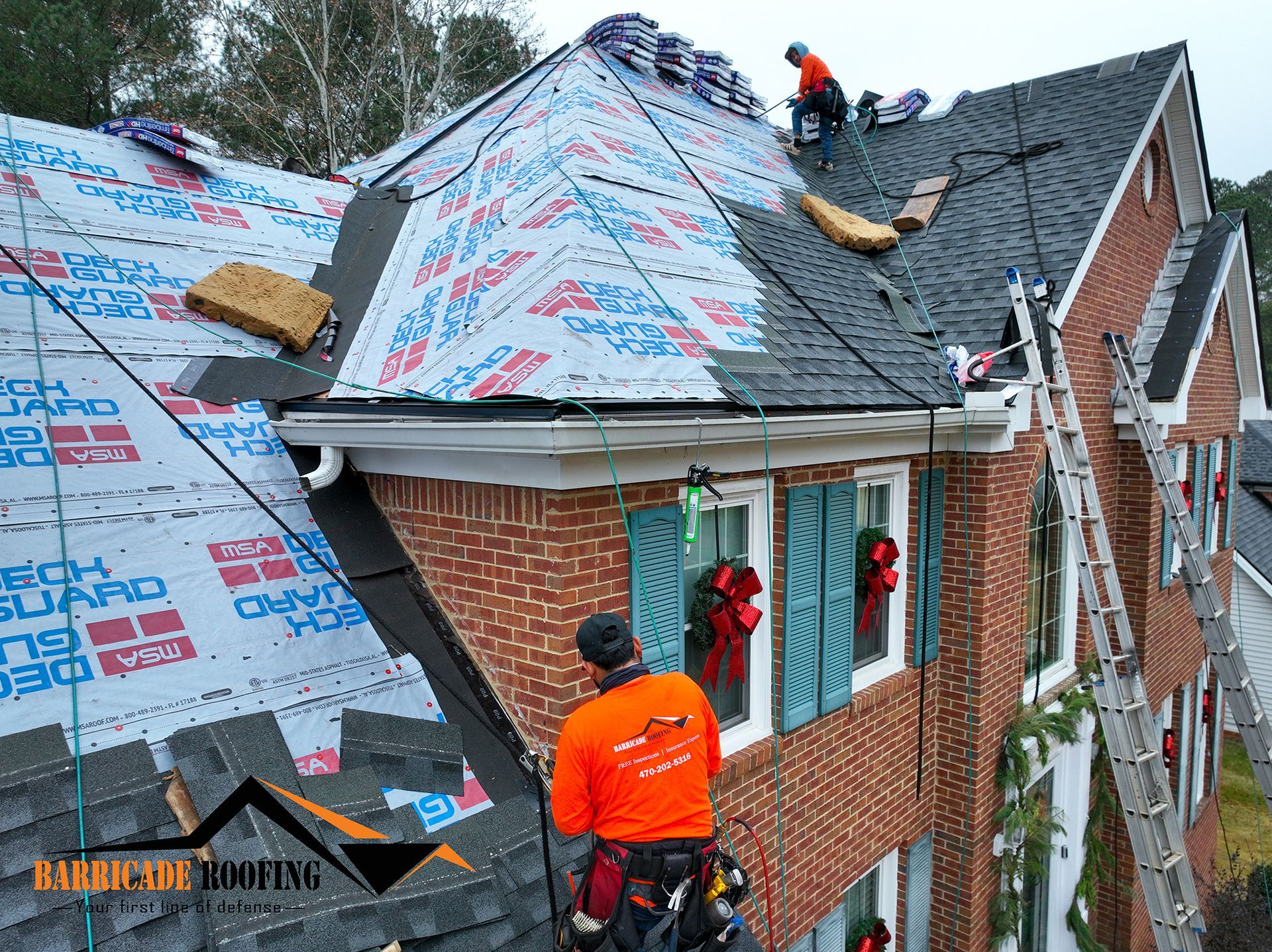 Roofers working on a brick house; one is harnessed, others on the roof.