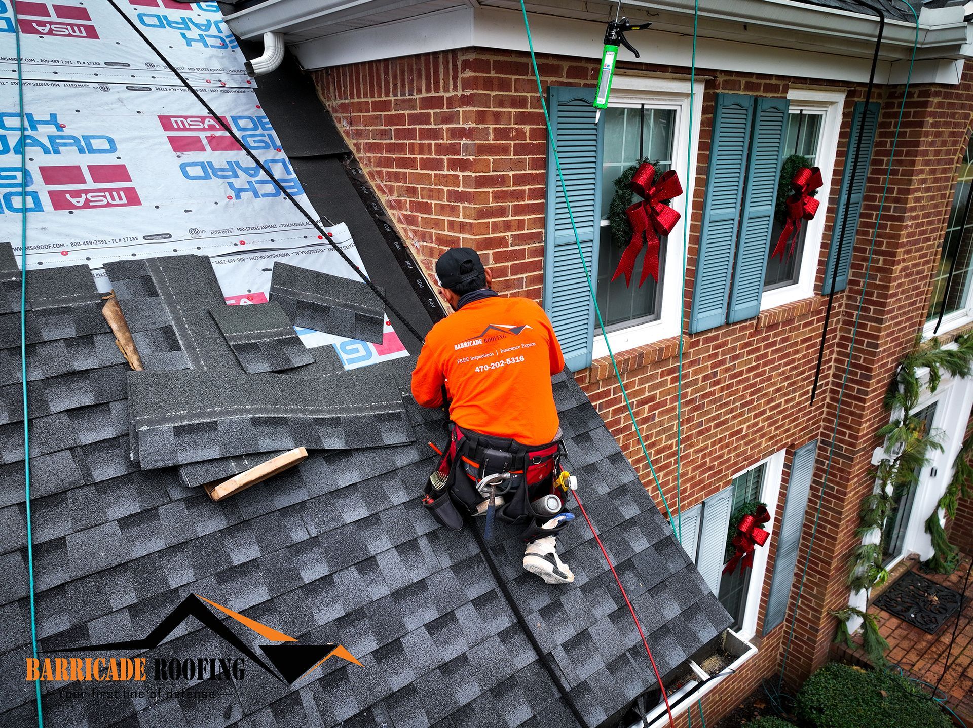 Roofer in orange shirt and harness works on a dark shingle roof, near a brick house with Christmas decorations.