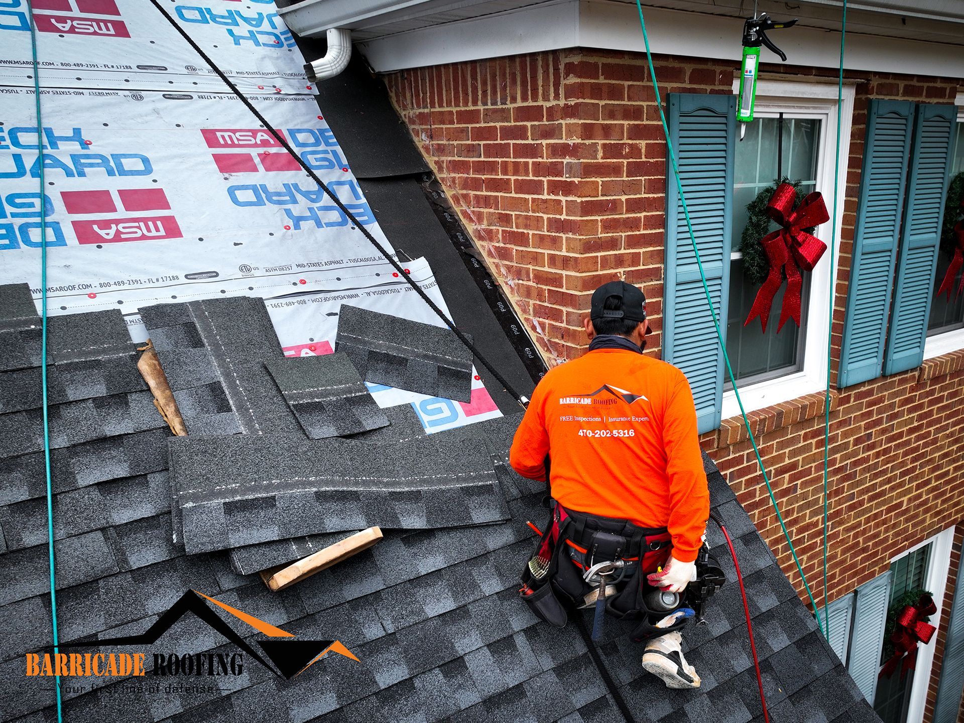 Roofer in orange shirt installing shingles on a house roof. Safety gear visible, blue shutters and wreath in background.
