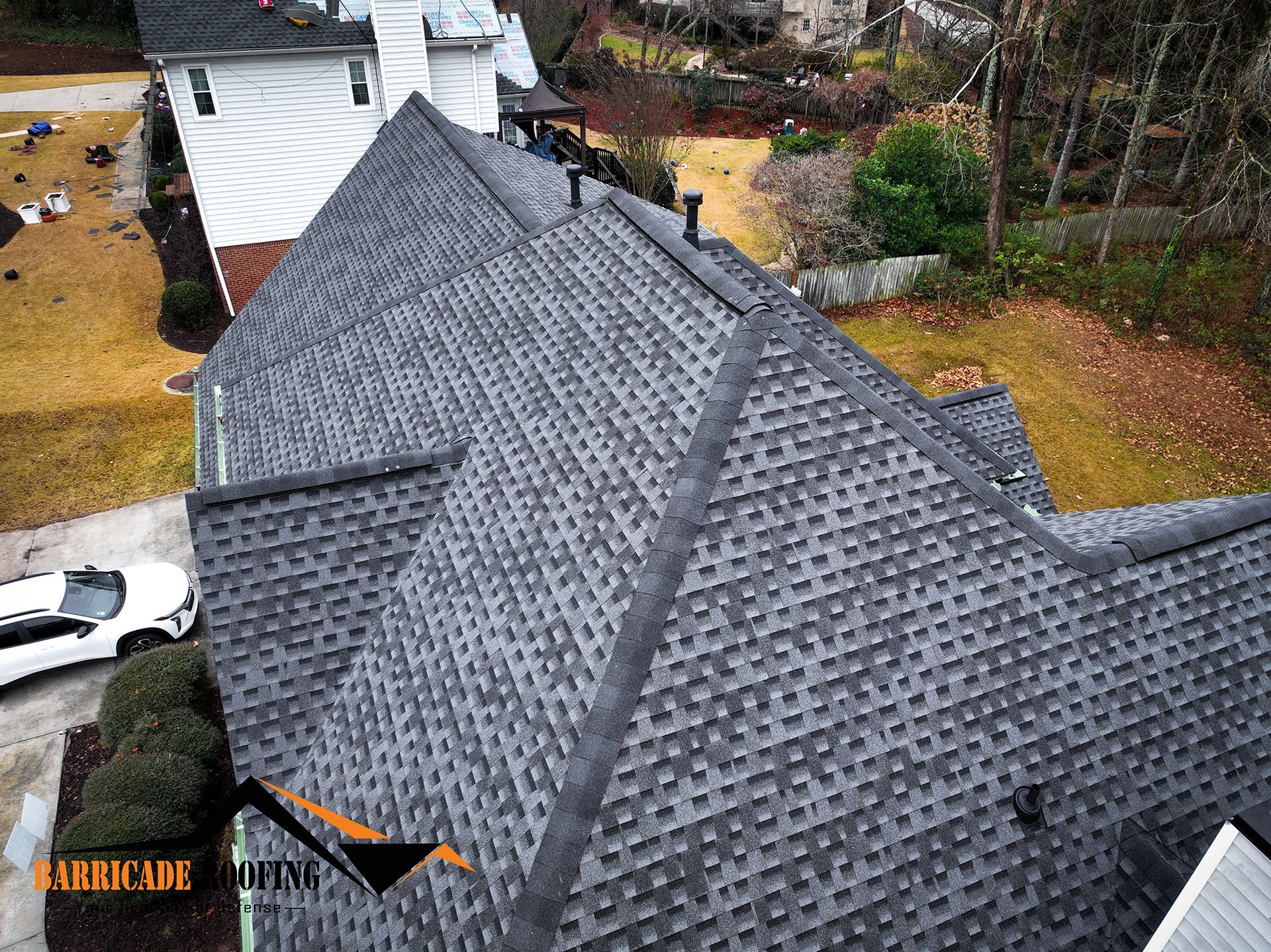 Overhead view of a house with a gray asphalt shingle roof. White car parked in the driveway. Trees and lawn in the background.