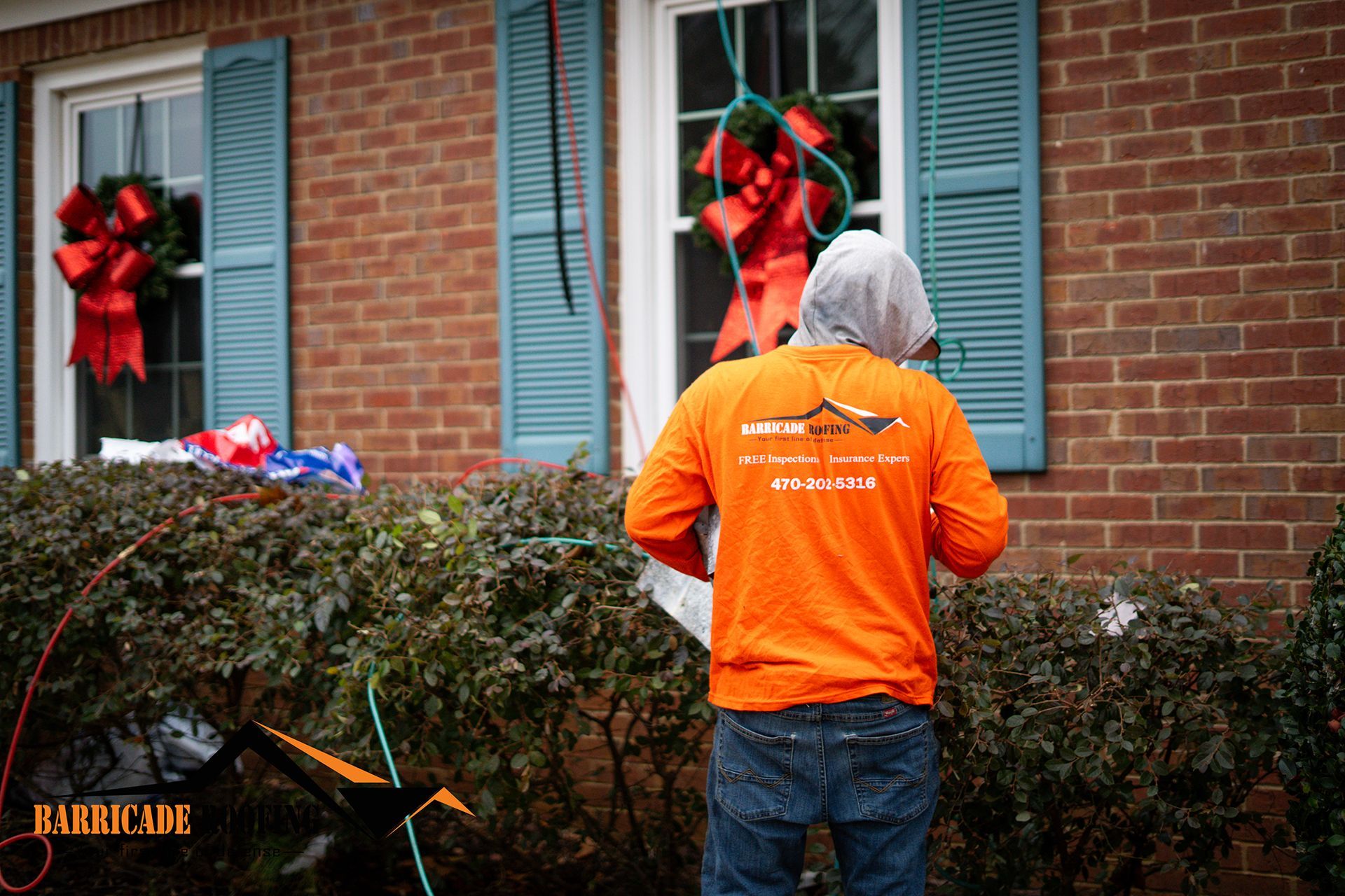 Person in orange shirt near a brick house with blue shutters, a wreath, and a red bow.