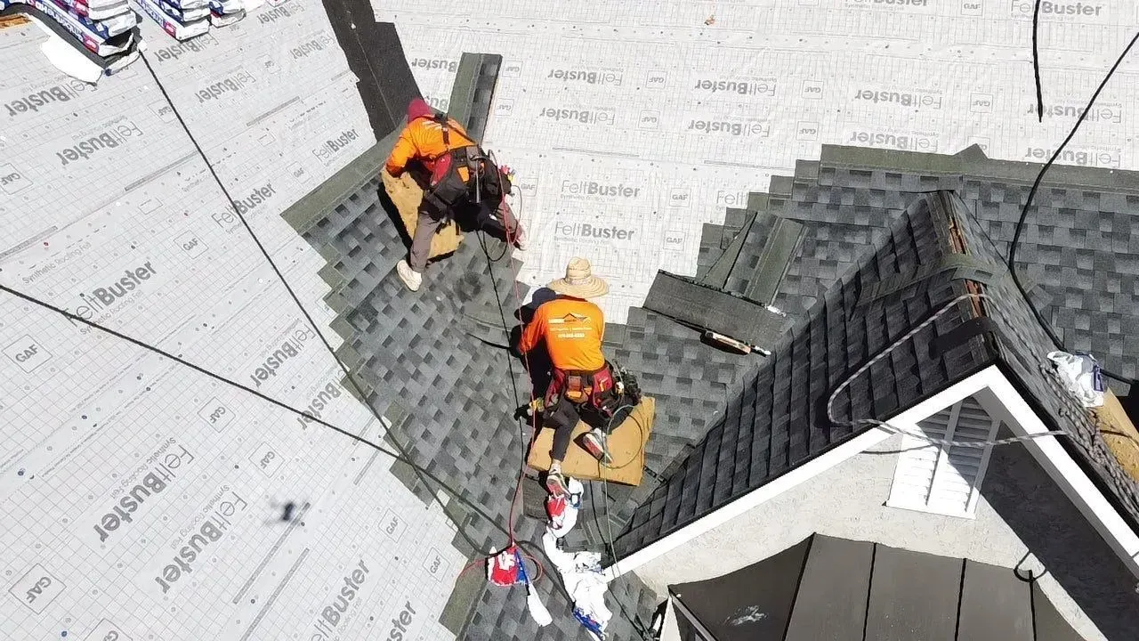 Two roofers installing shingles on a house roof. They wear safety gear and are working with tools.