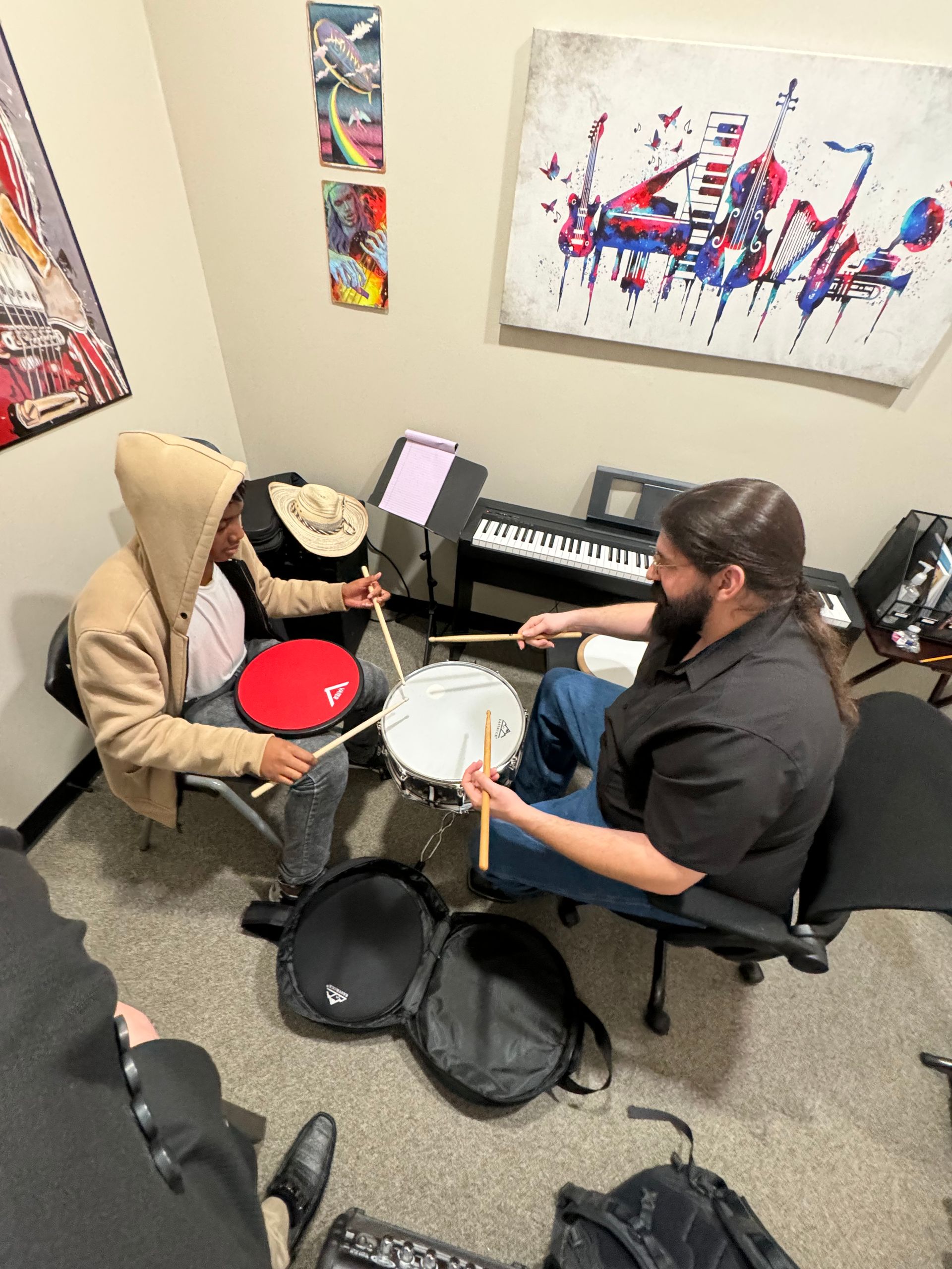 A man is teaching another man how to play drums in a room.