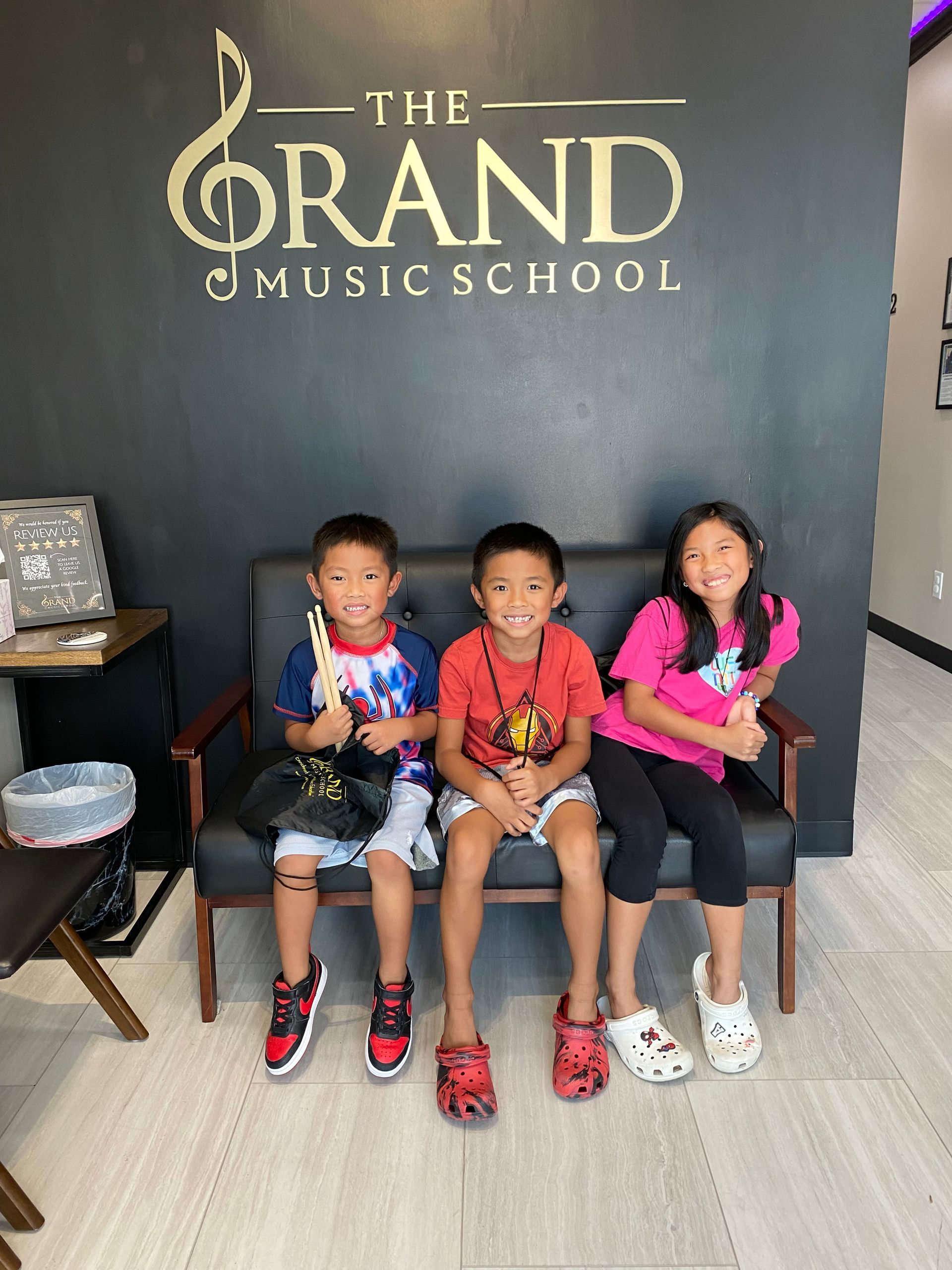 Three children are sitting on a couch in front of a sign for the grand music school