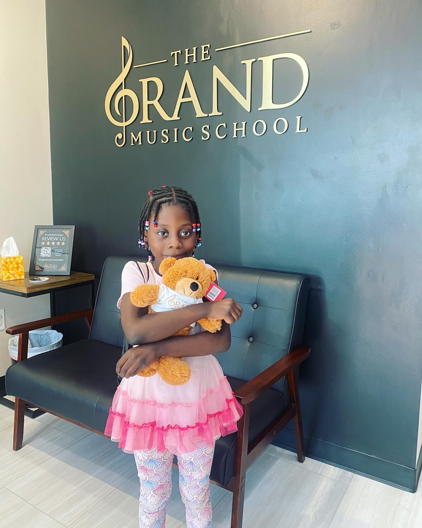 A little girl is holding a teddy bear in front of a grand music school sign.