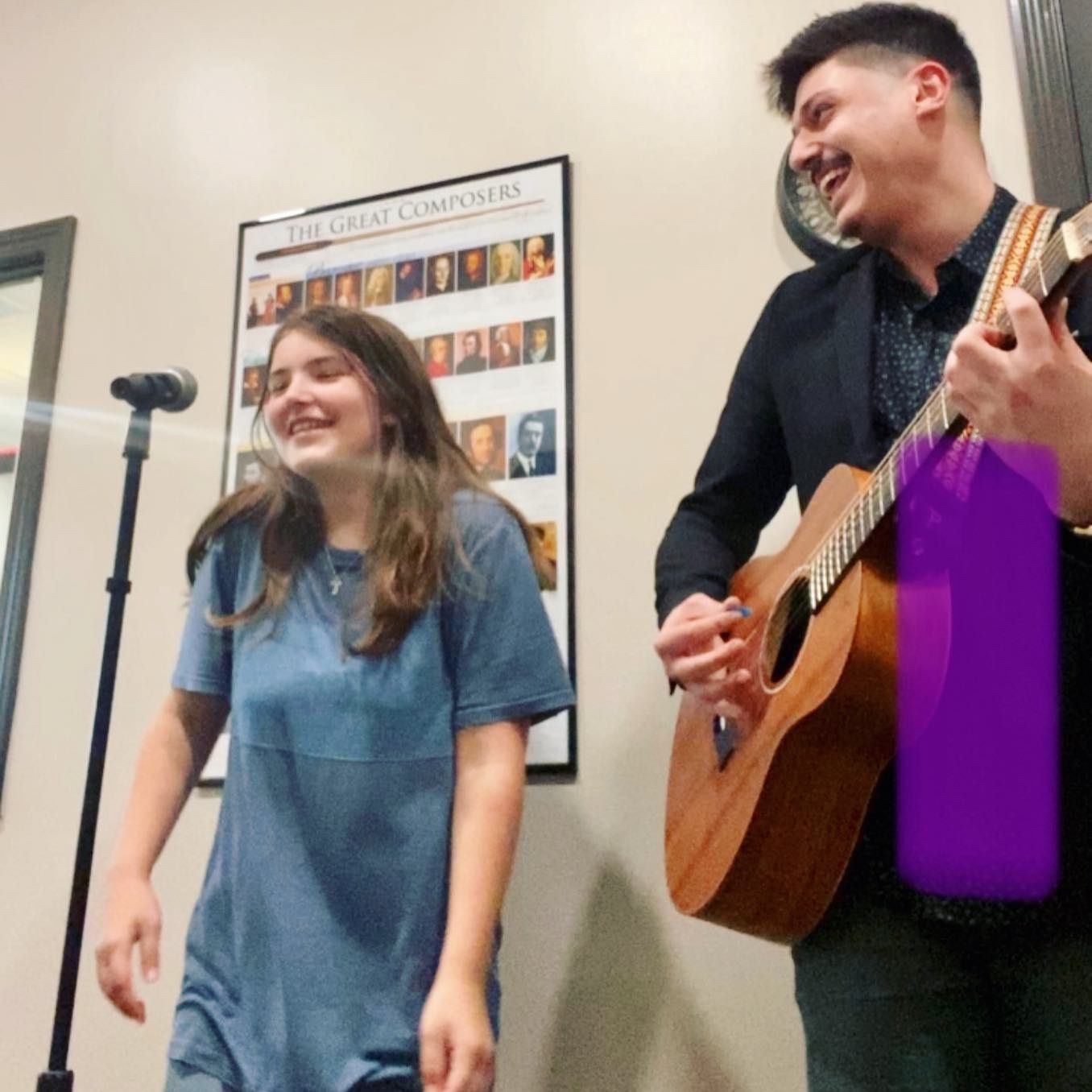 A man playing a guitar next to a woman in front of a poster that says the great composers