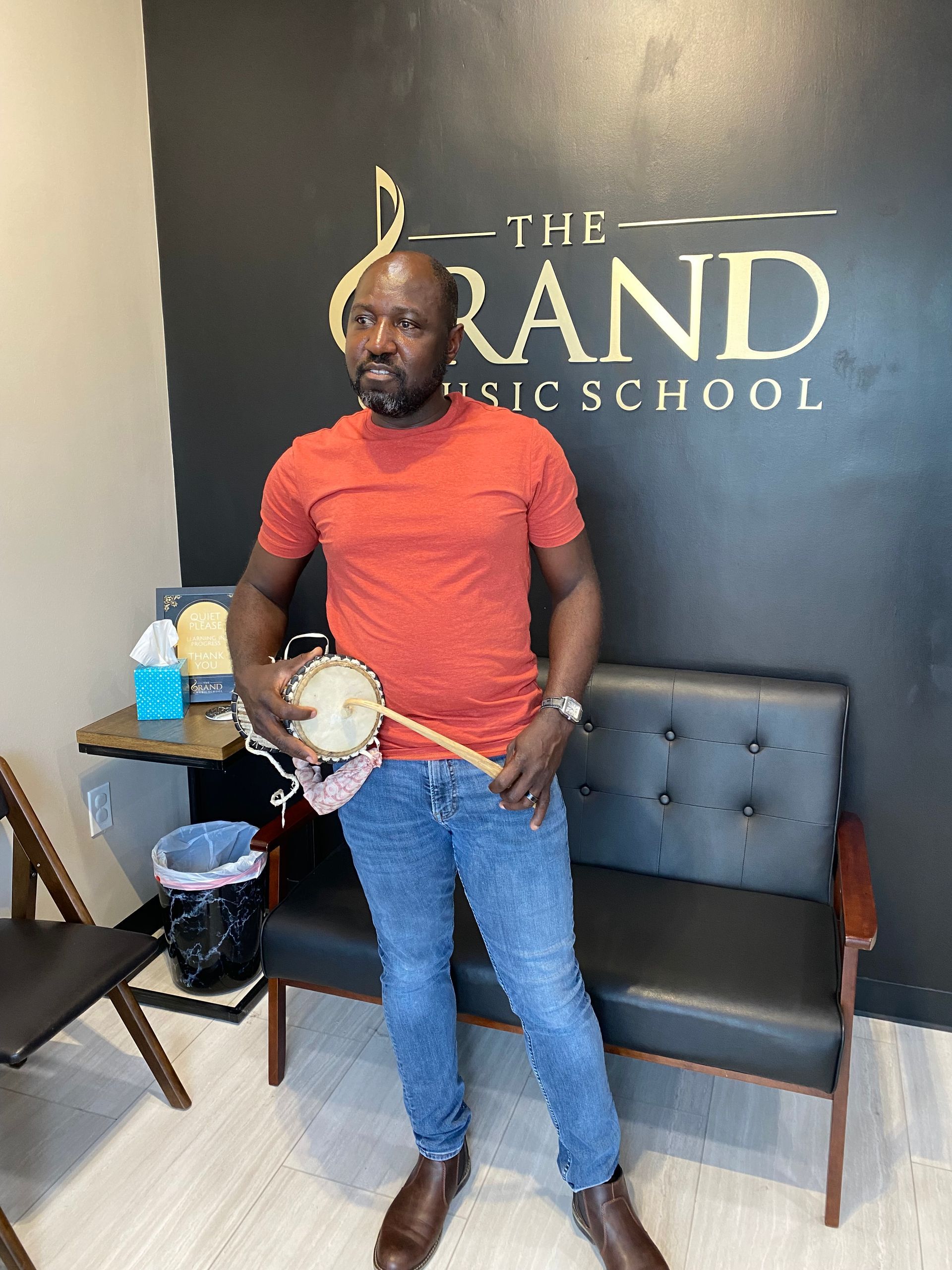 A man is holding a drum in front of a sign that says the grand music school.