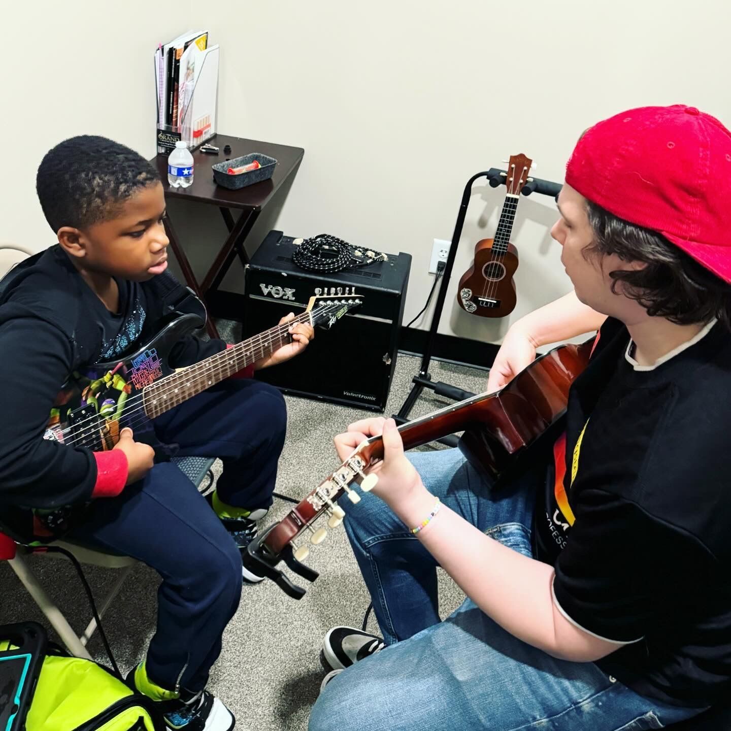 A man in a red hat is teaching a young boy how to play a guitar