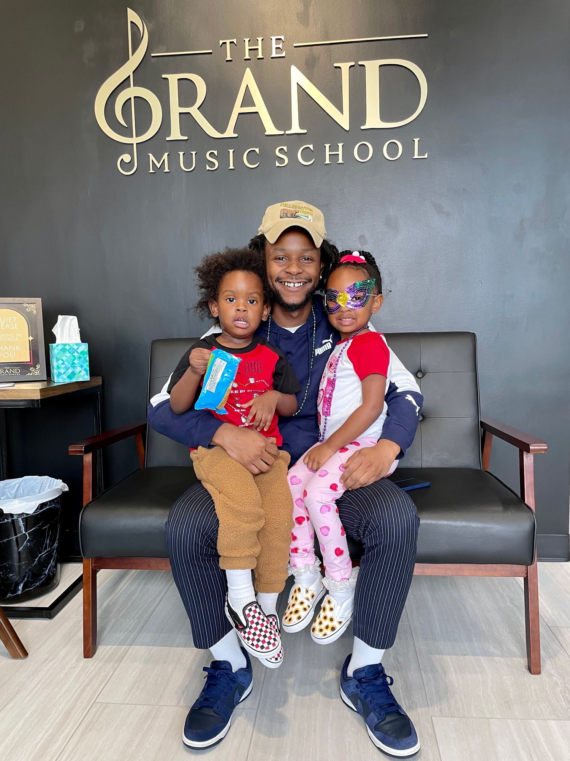 A man is sitting on a couch with two children in front of a sign for the grand music school.