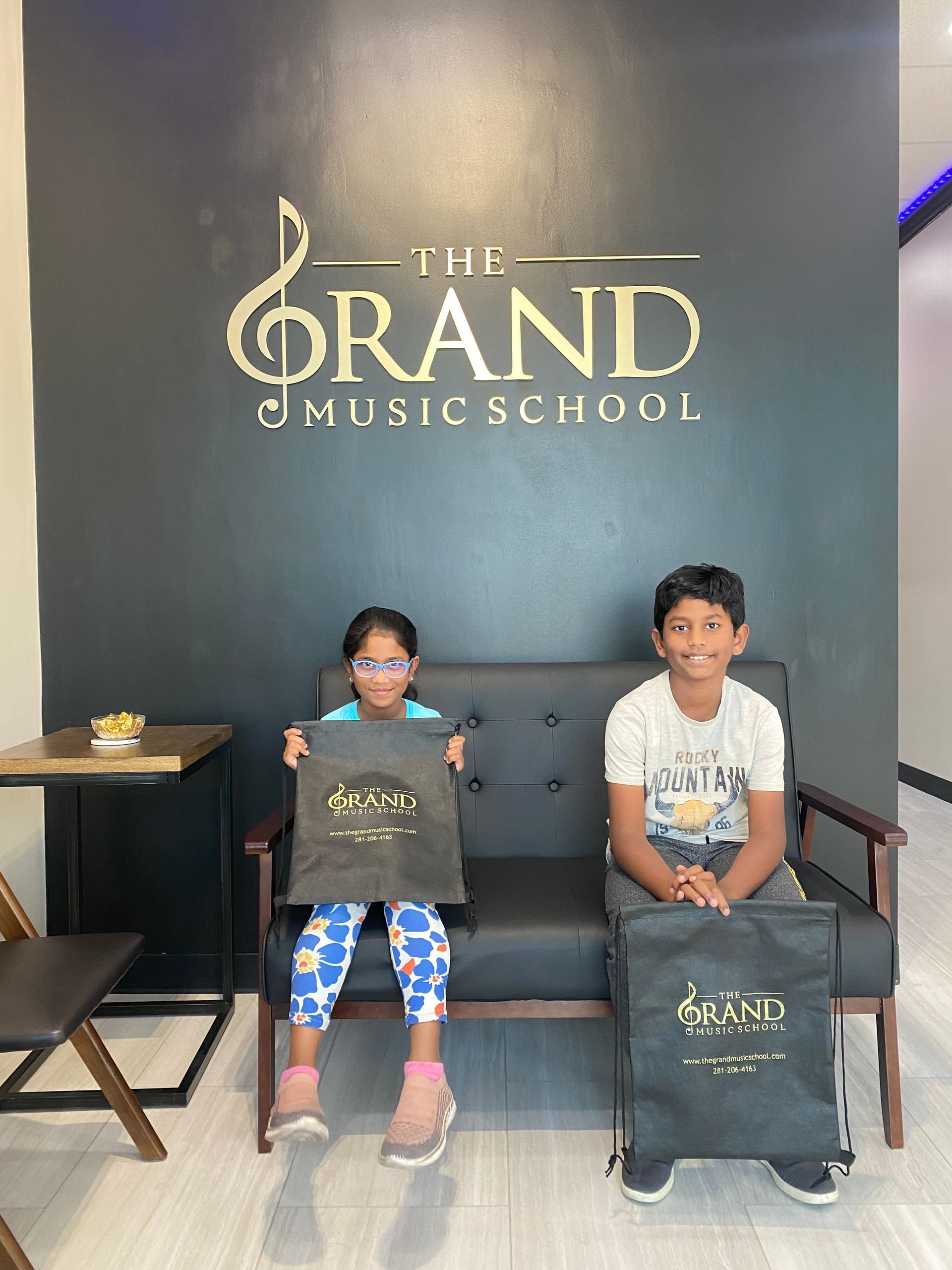 A boy and a girl are sitting on a couch in front of a wall that says the grand music school.
