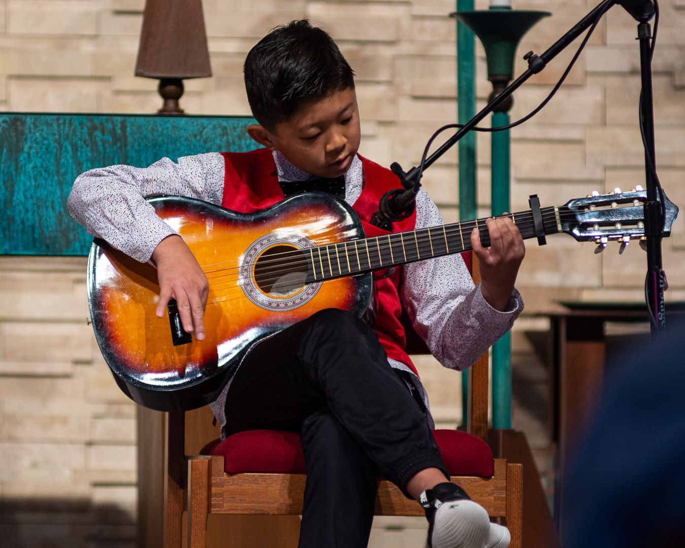 A young boy is sitting in a chair playing a guitar in front of a microphone.