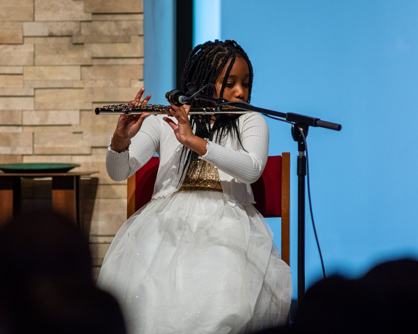 A young girl in a white dress is playing a flute in front of a microphone.
