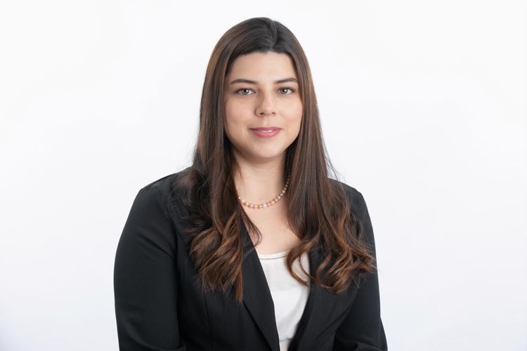 Woman with brown hair wearing a black blazer, white shirt, and a pearl necklace, smiling against a white background.