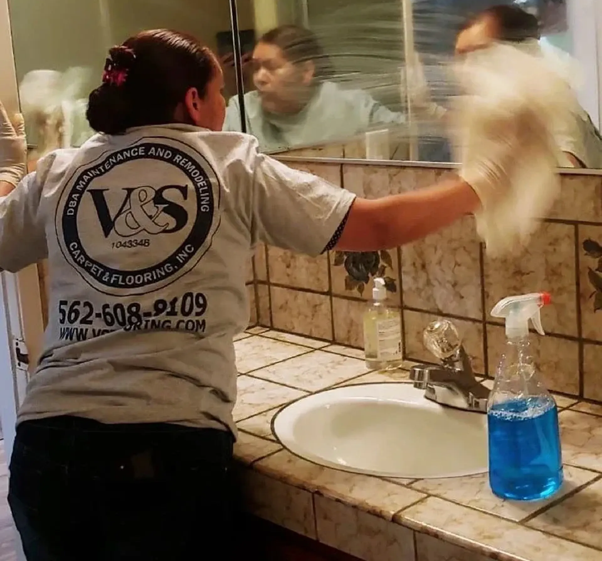 Person cleaning bathroom mirror with cleaning solution and cloth; sink visible.