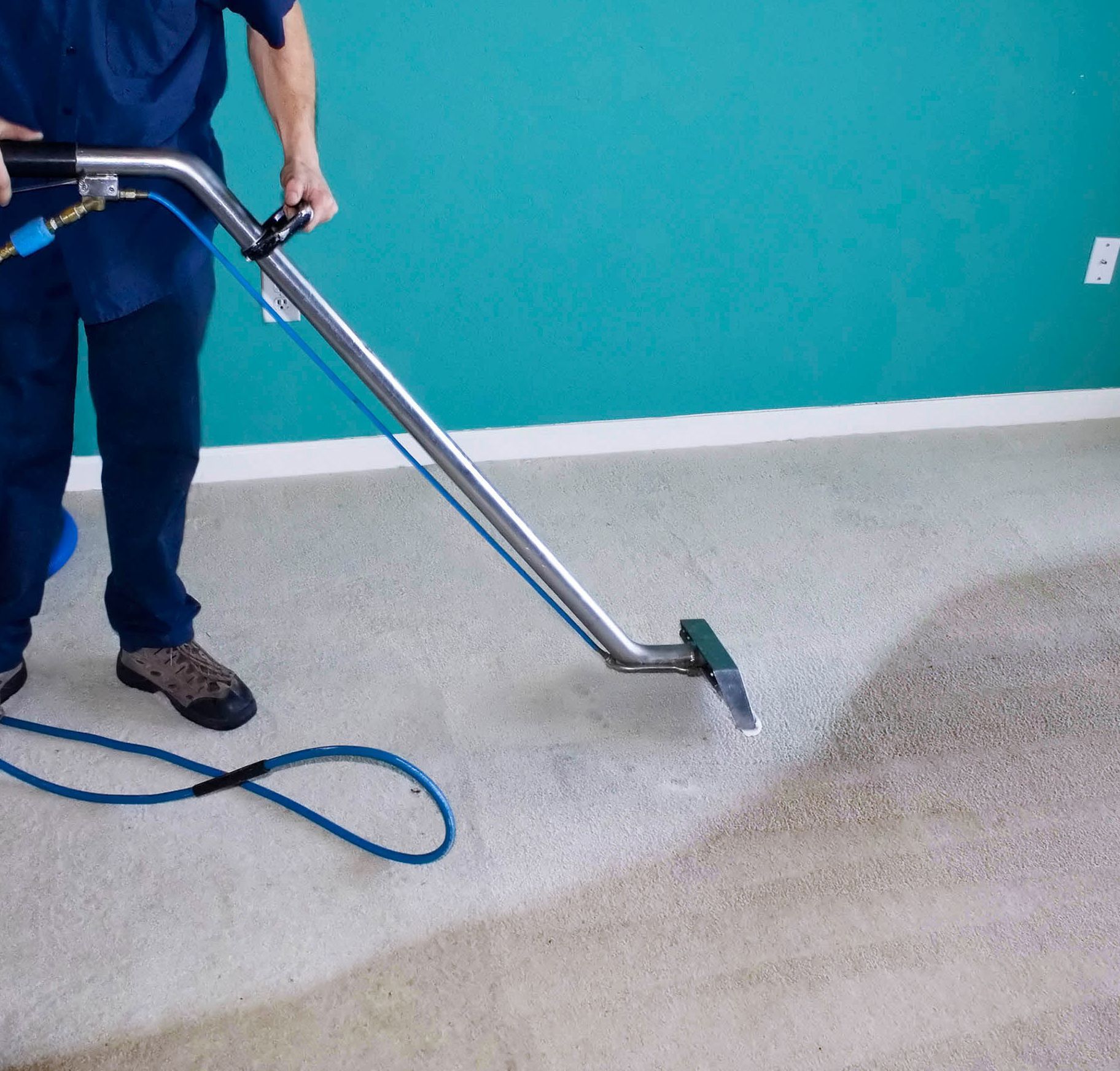 Person in blue uniform cleaning a carpet with a steam cleaner.