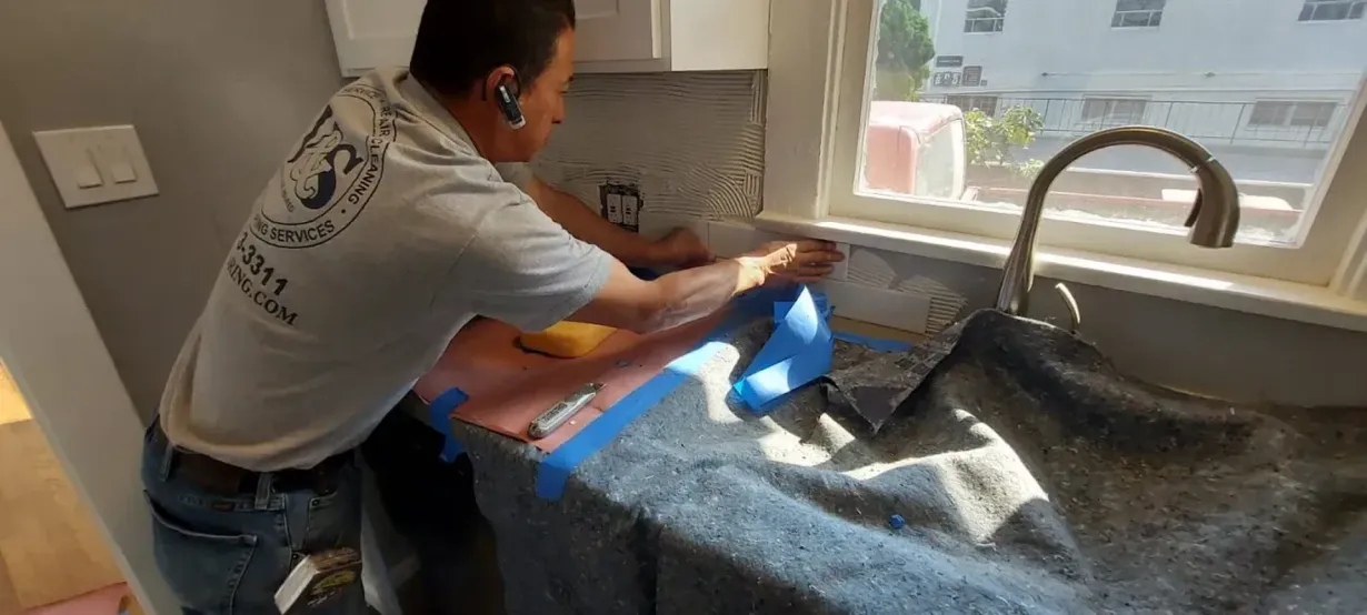 A person is installing backsplash in a kitchen. Blue tape is on the countertop. A sink and window are also in the scene.