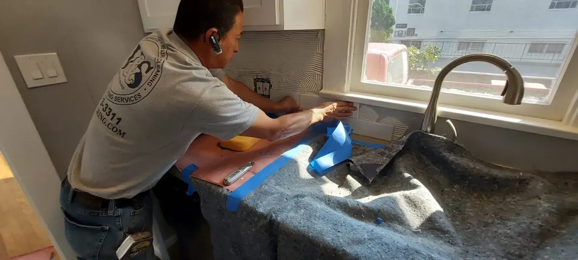 A person is installing backsplash in a kitchen. Blue tape is on the countertop. A sink and window are also in the scene.
