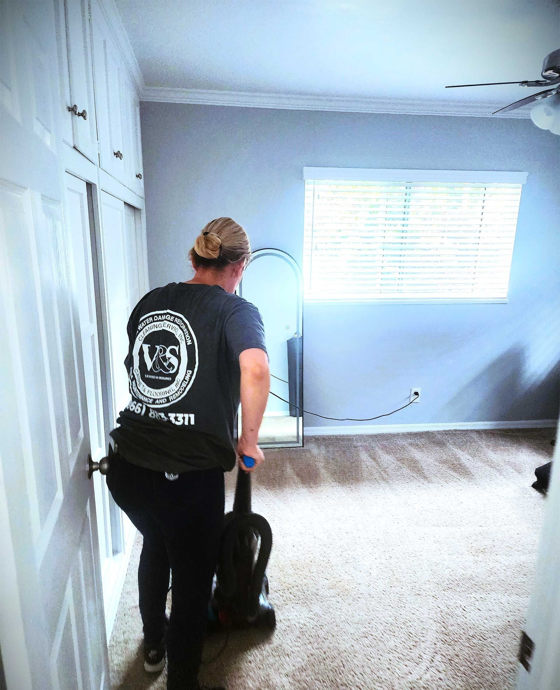 Person vacuuming carpeted room; neutral colors, sunlight through window, door ajar.