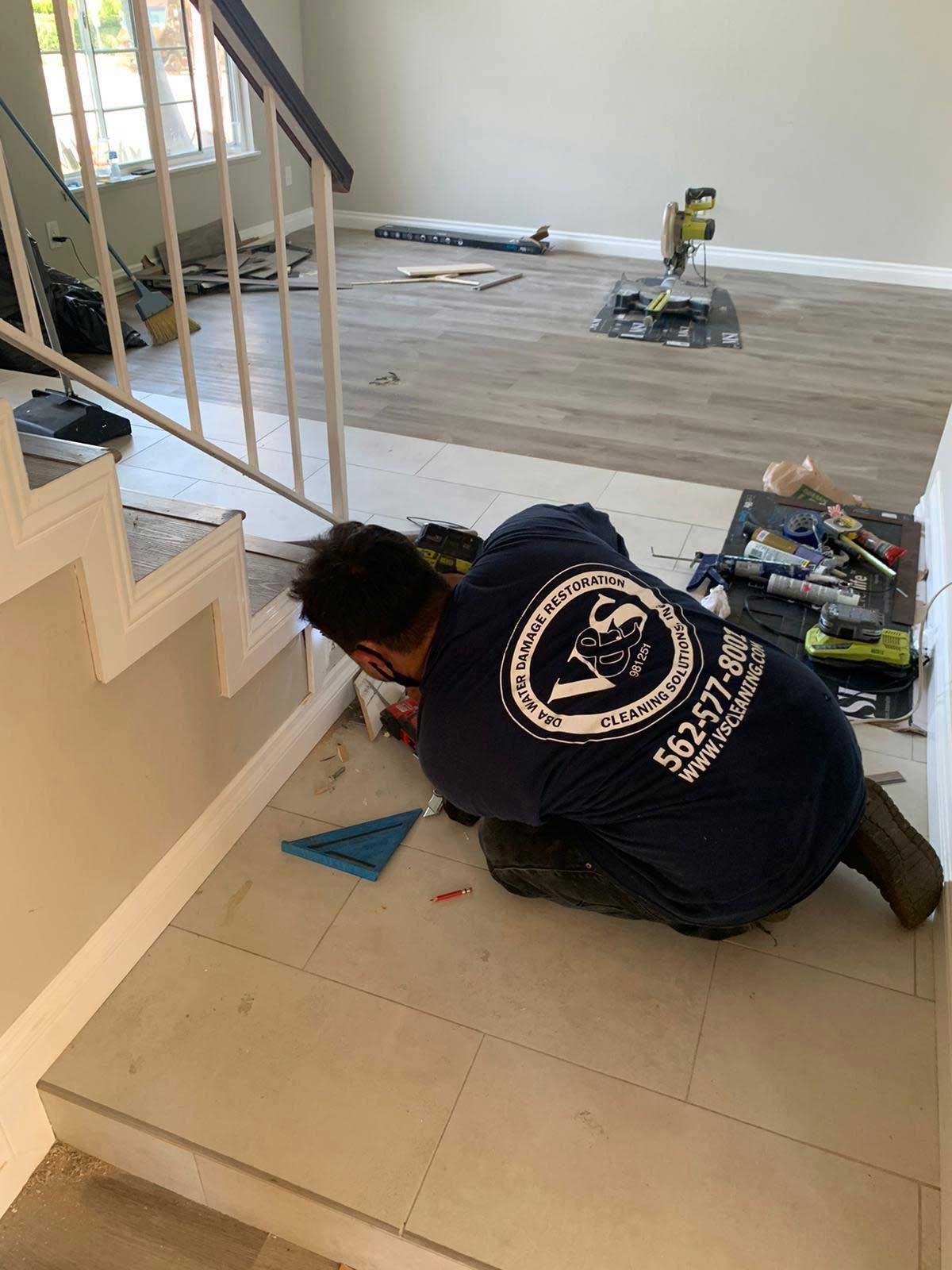 A person kneeling, installing baseboard tile. Tools and stairs are visible.