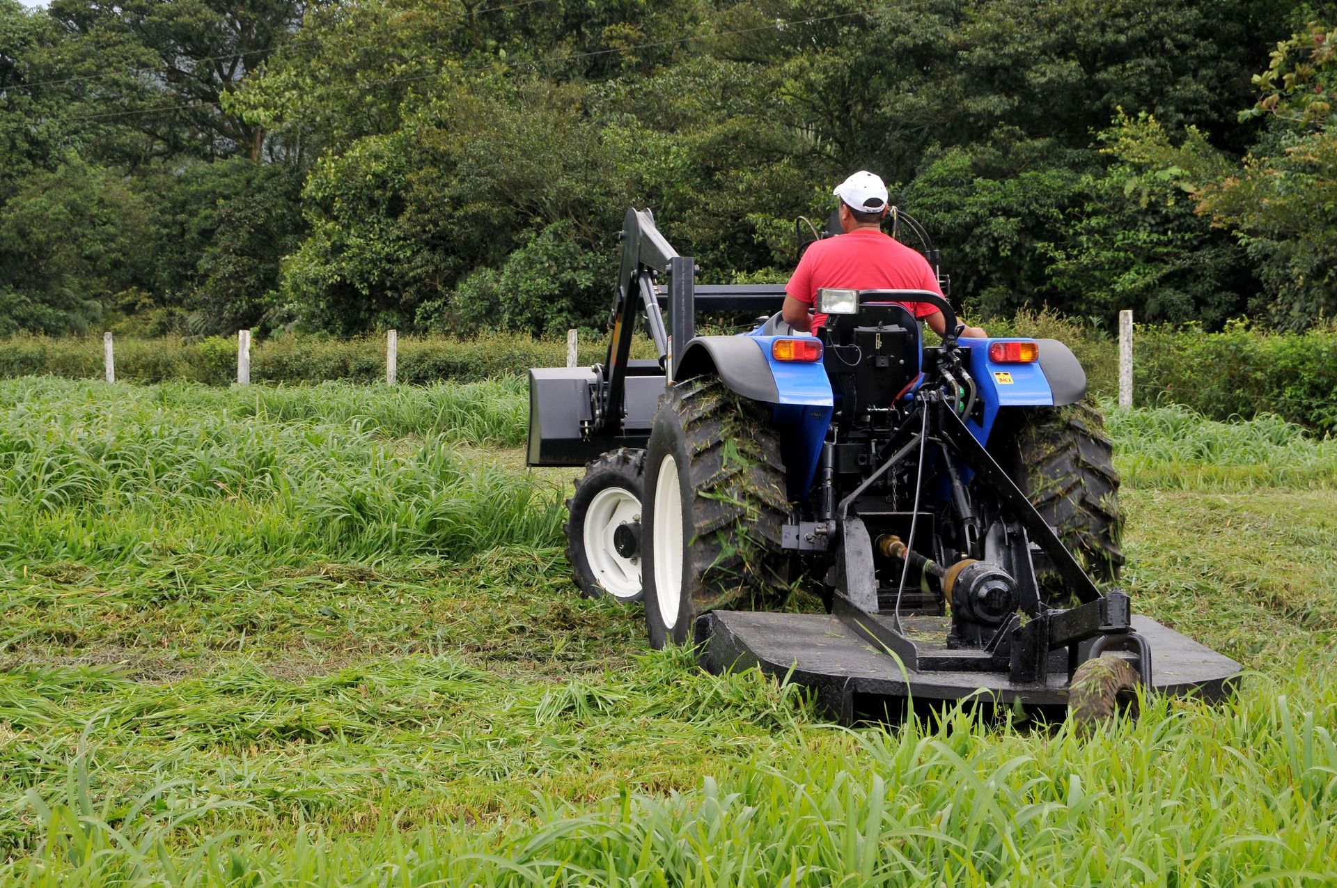 Man on tractor mowing a field of grass. Blue tractor, red shirt, white hat. Green field, trees in background.