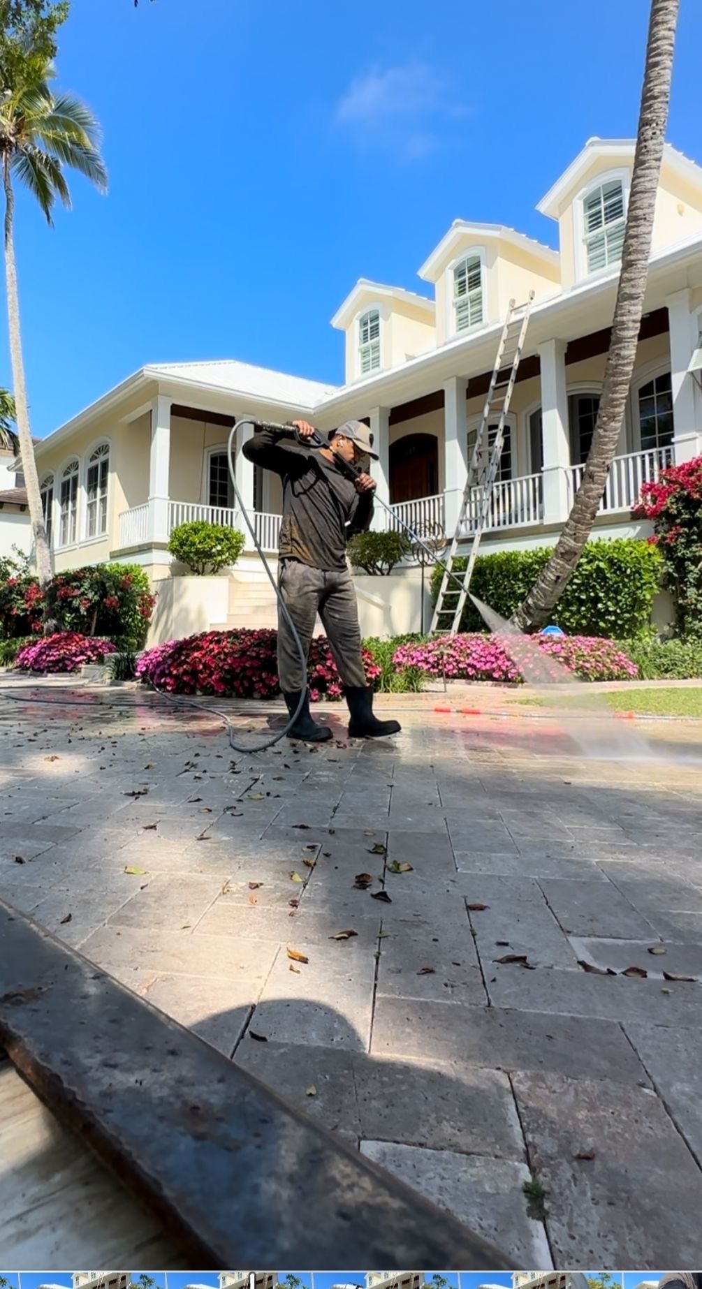 Person standing on a paved walkway in front of a large white house with columns and pink flowers.