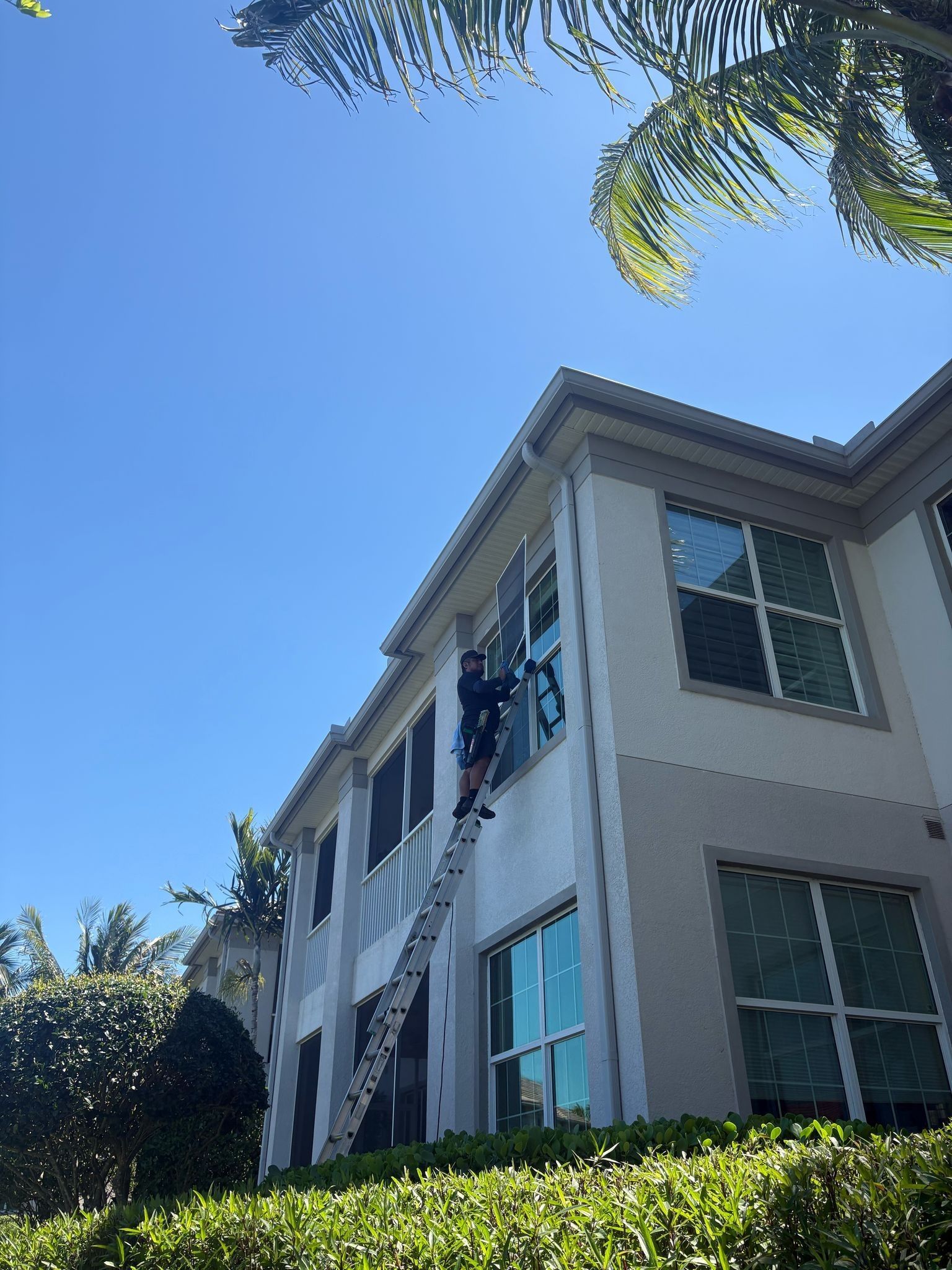 Two-story beige house with blue windows, seen from below against a clear blue sky and trees
