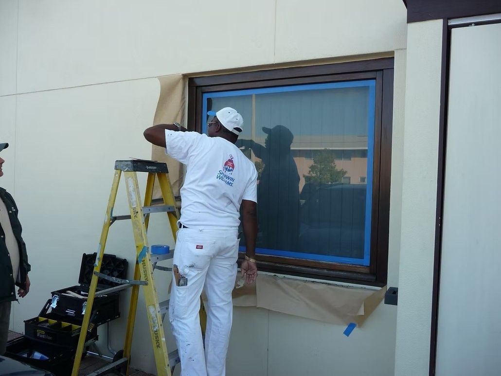 Worker painting a building wall beside a window, with a ladder and tools nearby.