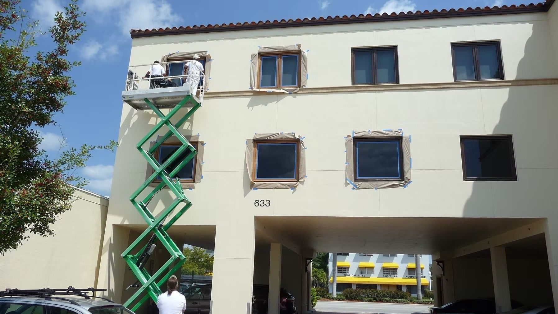 A worker on a green scissor lift repairs the upper left side of a cream-colored building.