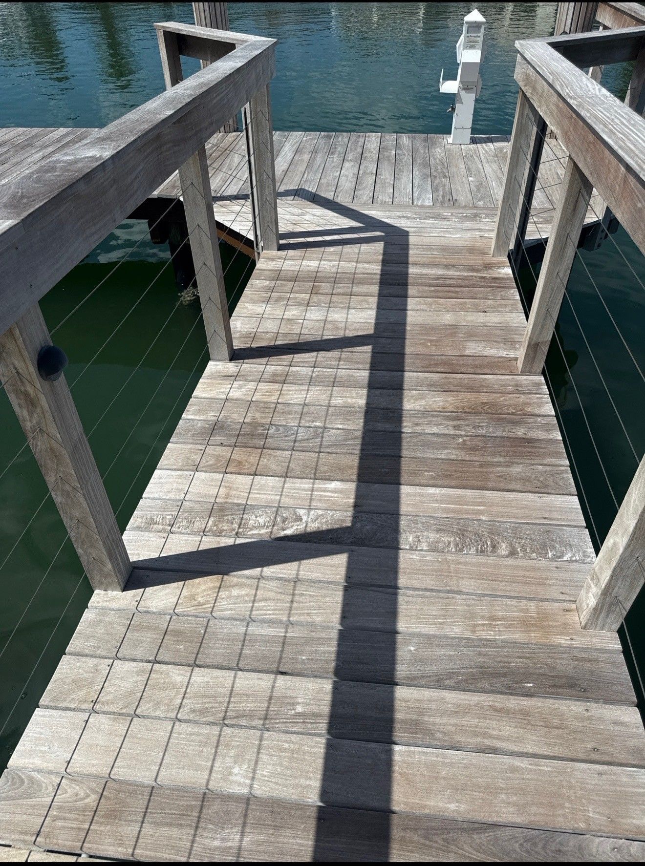 Wooden dock walkway with railings leading to blue water, casting strong shadows.