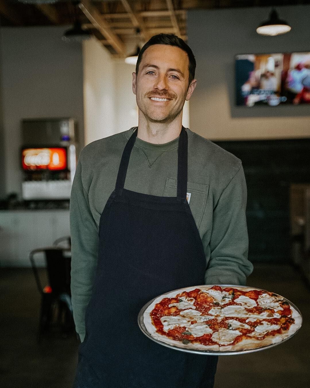 A man in an apron is holding a pizza in a restaurant.