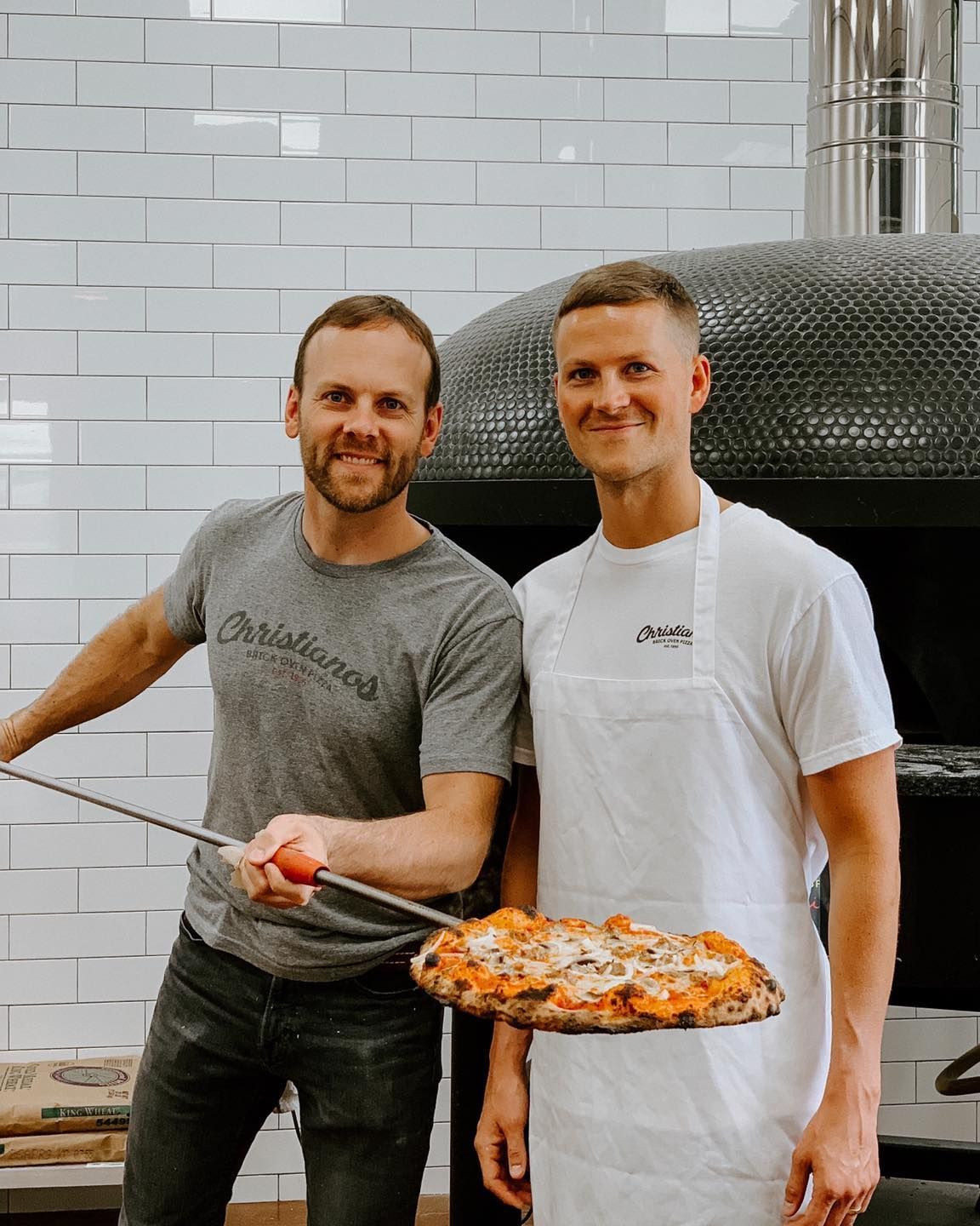 Two men are standing next to each other holding a pizza.
